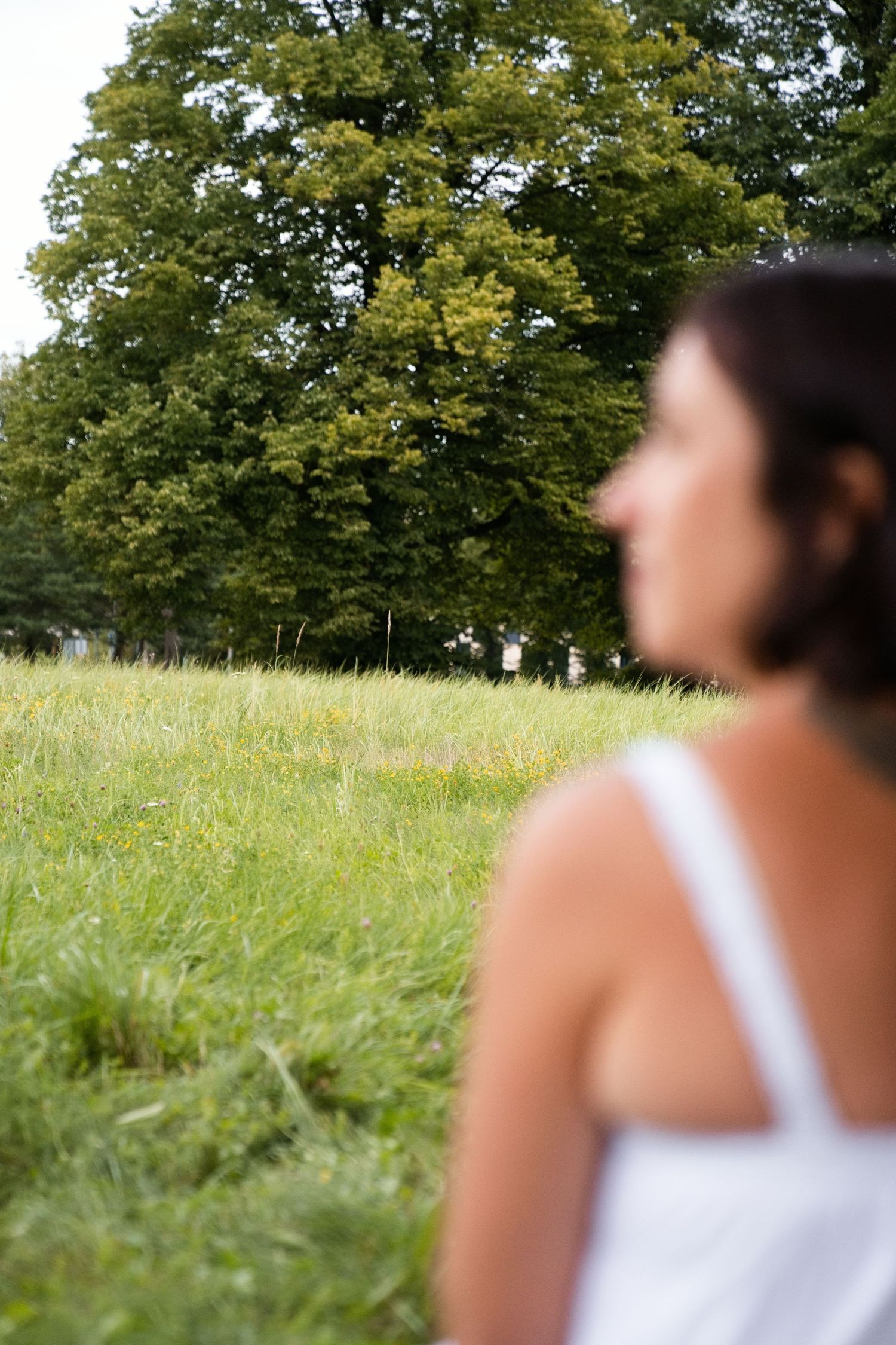 Woman in white tank top gazing at a lush green meadow with large trees in the background on a summer day.