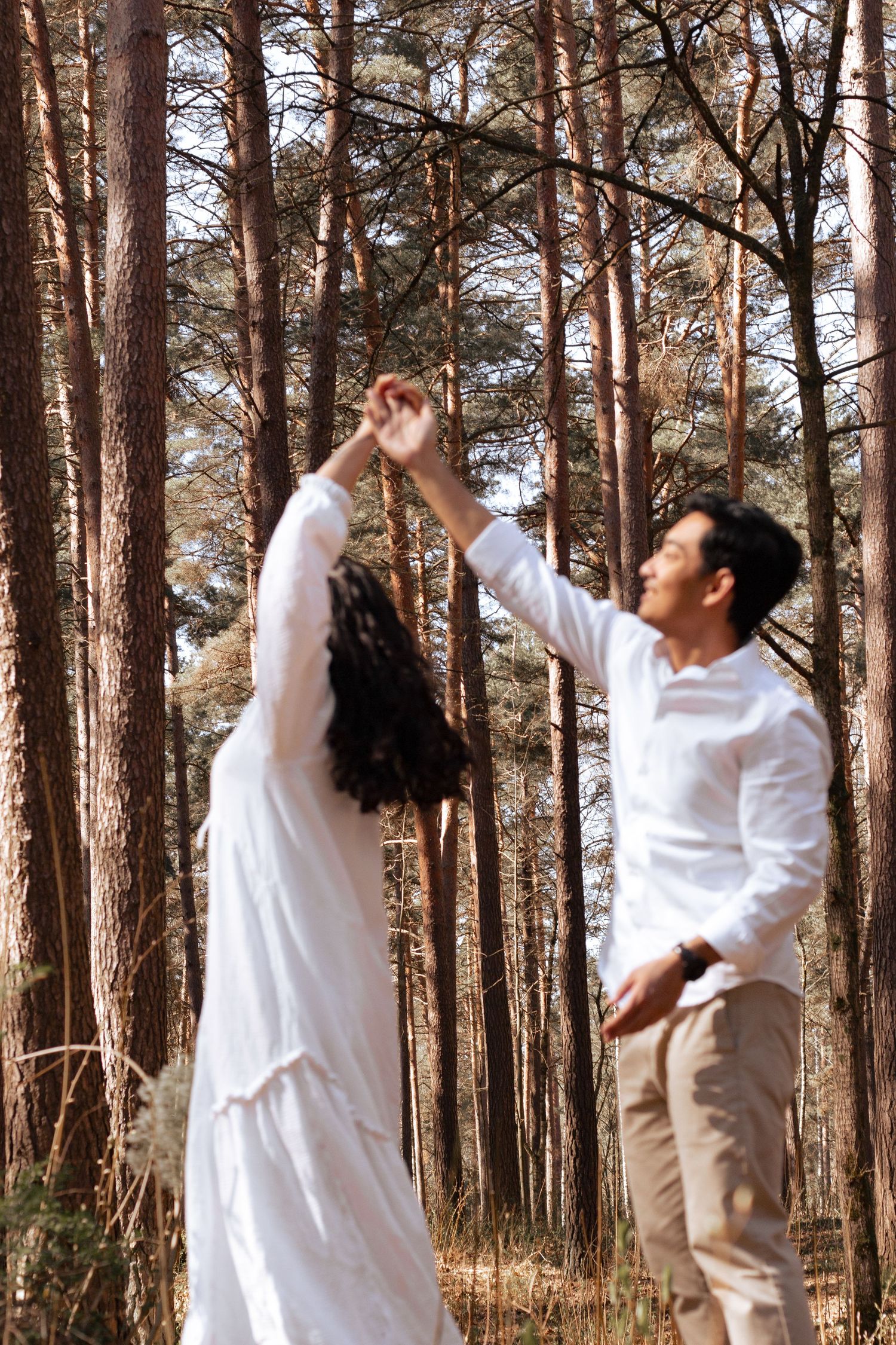 Couple dancing in a sunlit pine forest, woman in white dress with man in white shirt holding hands raised.