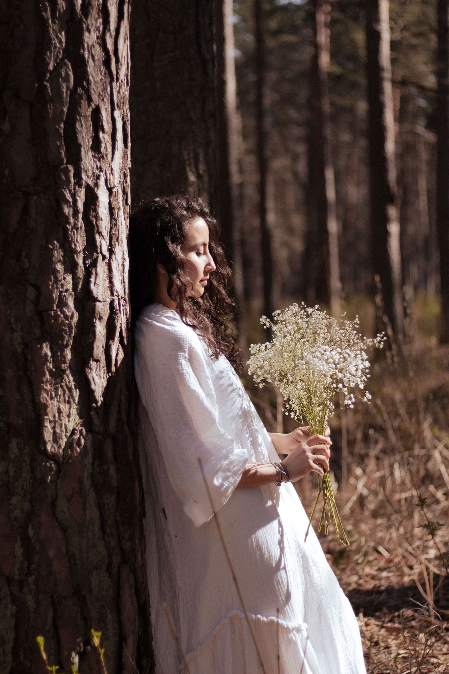 Woman in white dress holding wildflowers leaning against tree in sunlit forest.