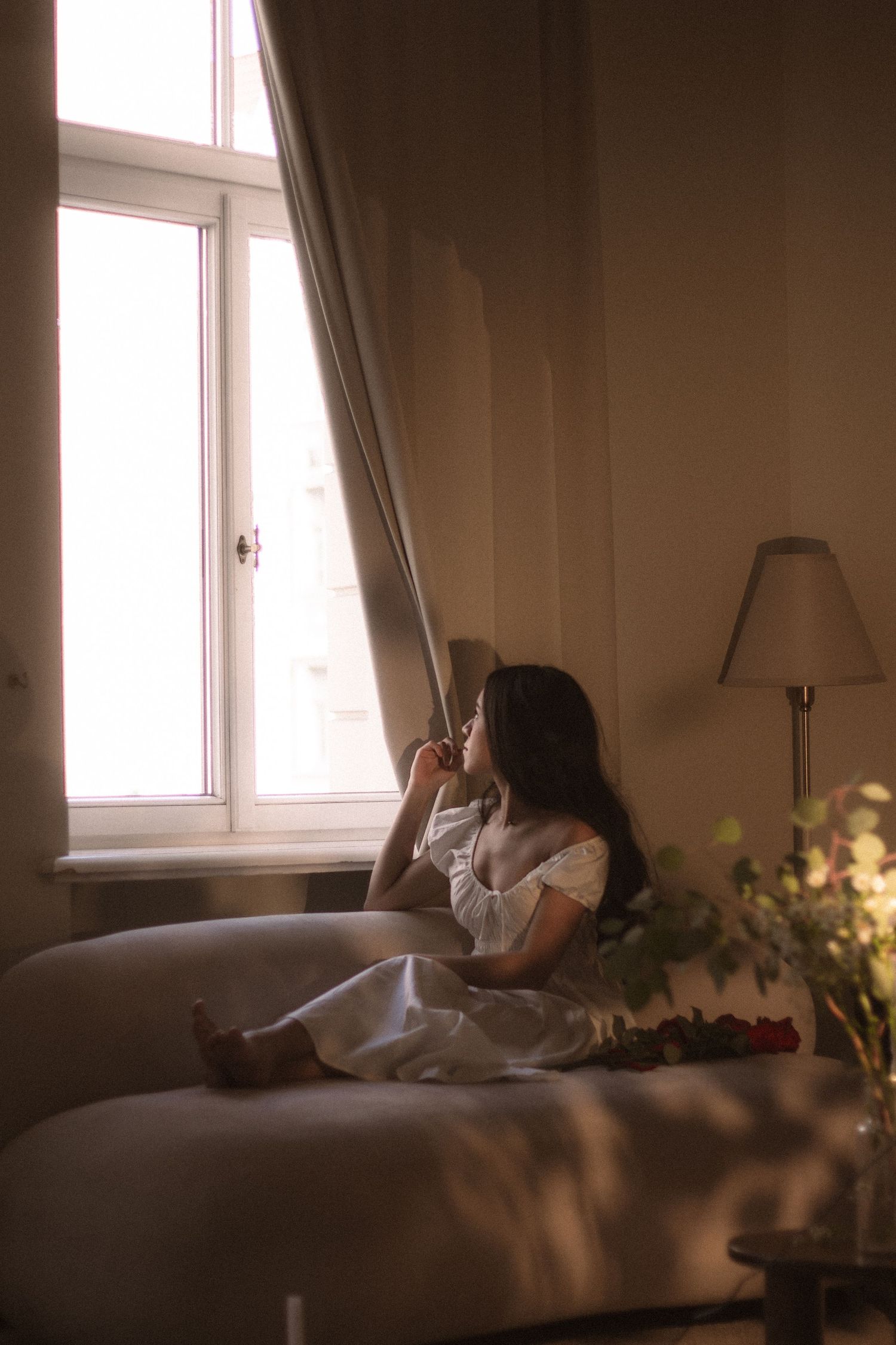 Young woman in a white dress sitting on a sofa gazing out a sunlit window in a warmly lit room.