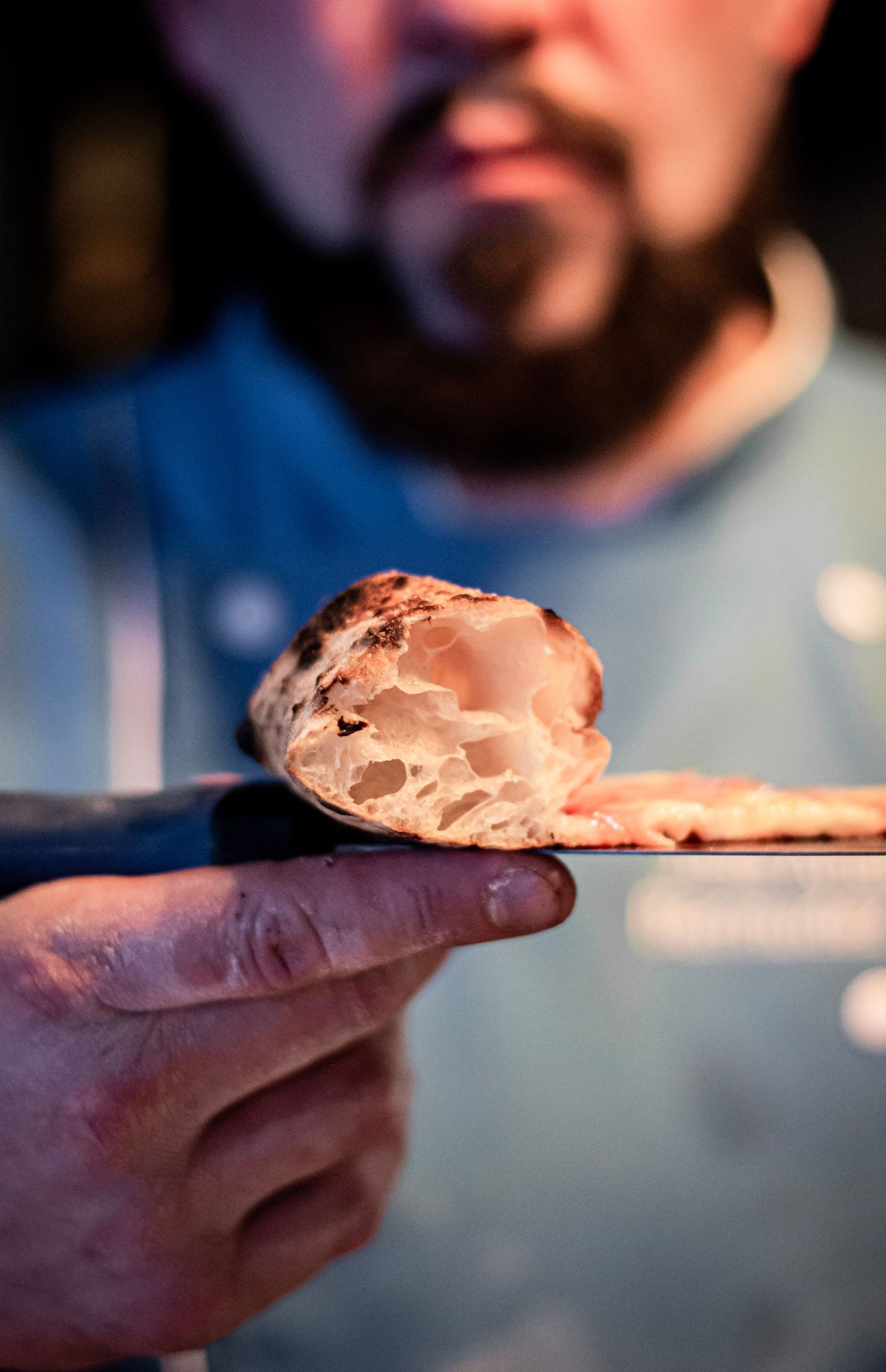 Bearded chef holding a slice of Neapolitan pizza crust showing airy open crumb structure.