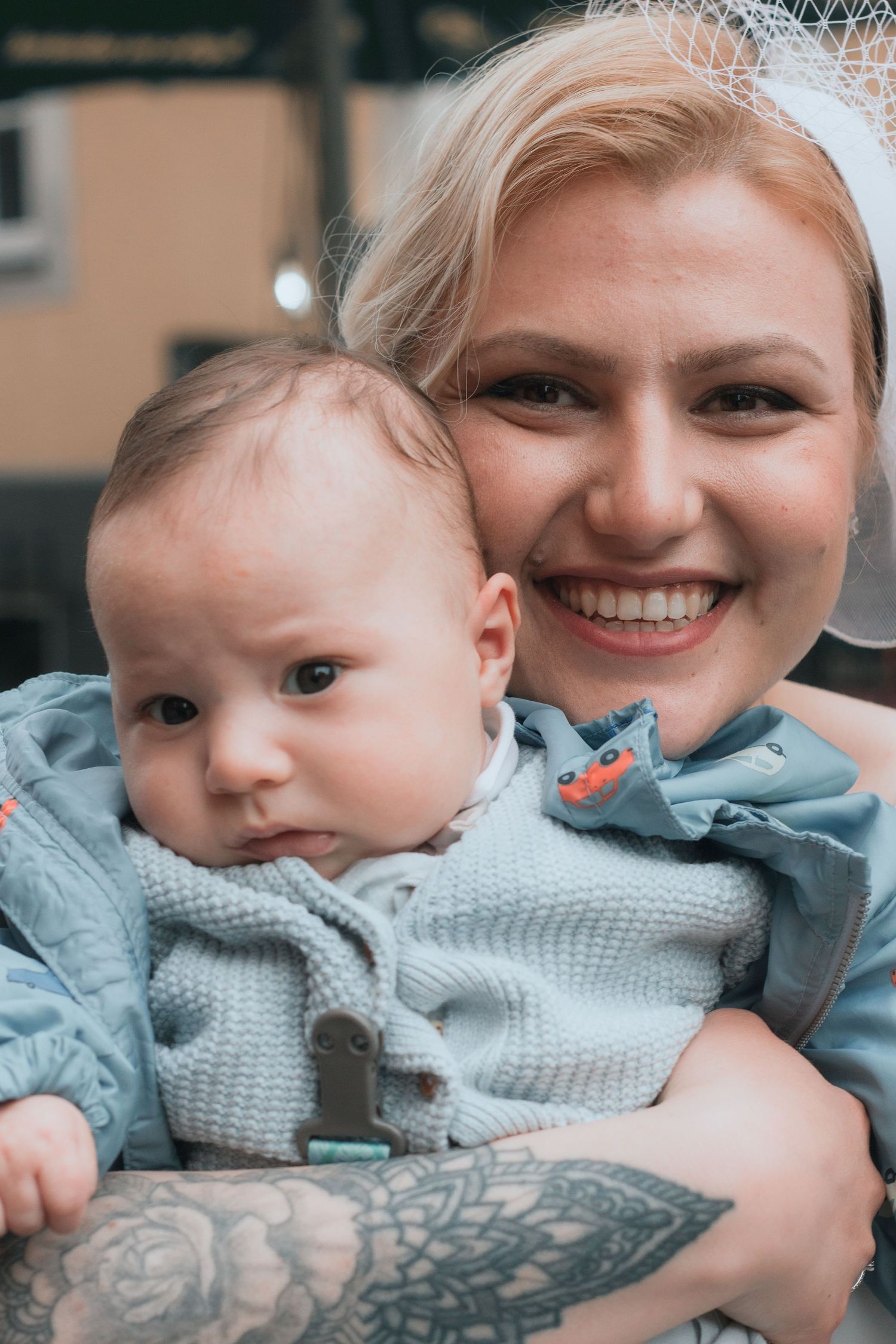 Smiling blonde woman with tattoos holding a cute baby dressed in light blue knit outfit.