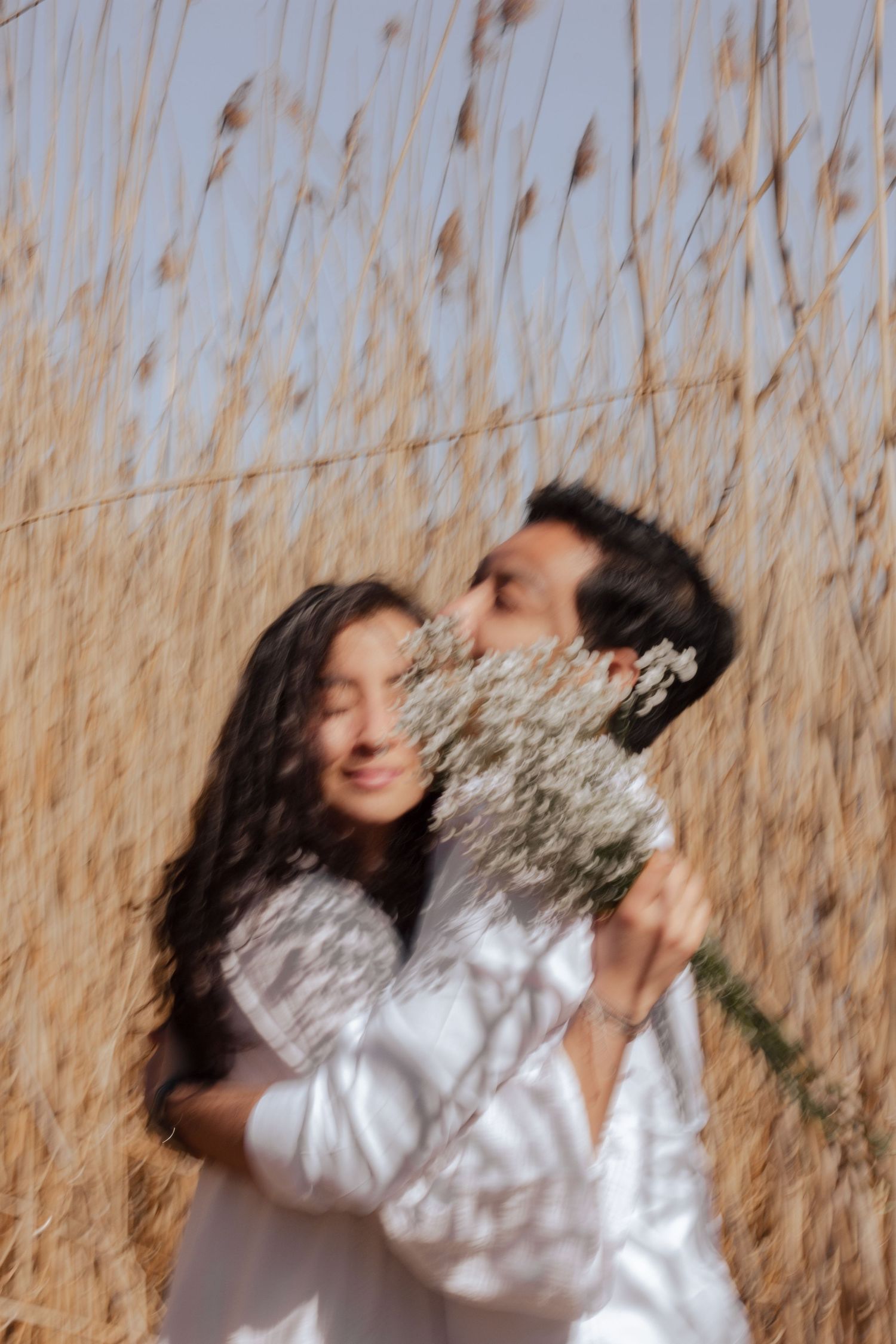 Romantic couple kissing behind bouquet of white wildflowers in golden grass field.