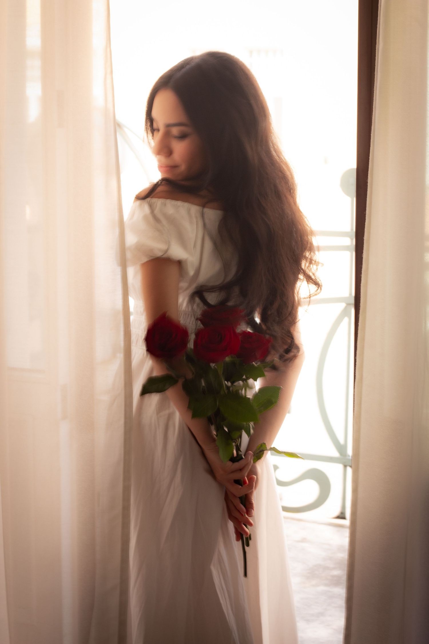 Young woman in white dress holding red roses standing in sunlit balcony doorway.