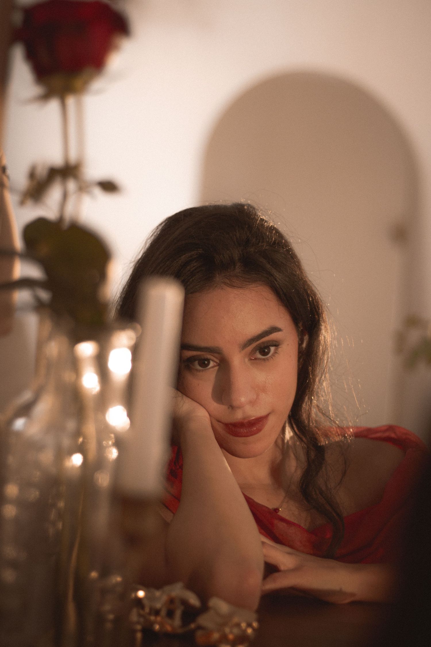 Young woman in red top resting chin on arms at a table with candles and a red rose nearby.
