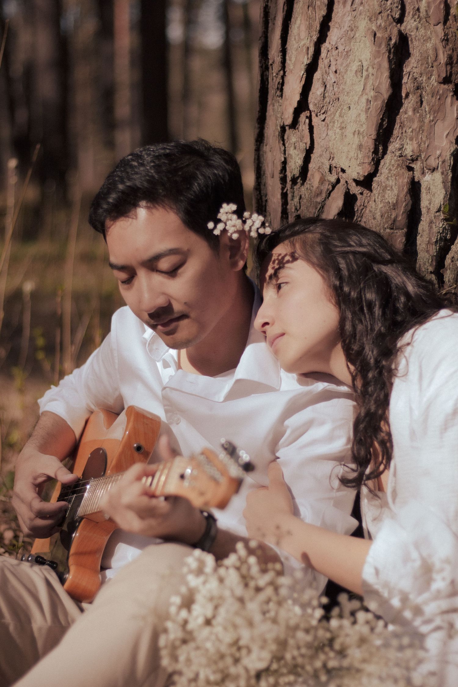 Couple in white shirts sitting against a tree, man playing ukulele while woman leans on his shoulder.