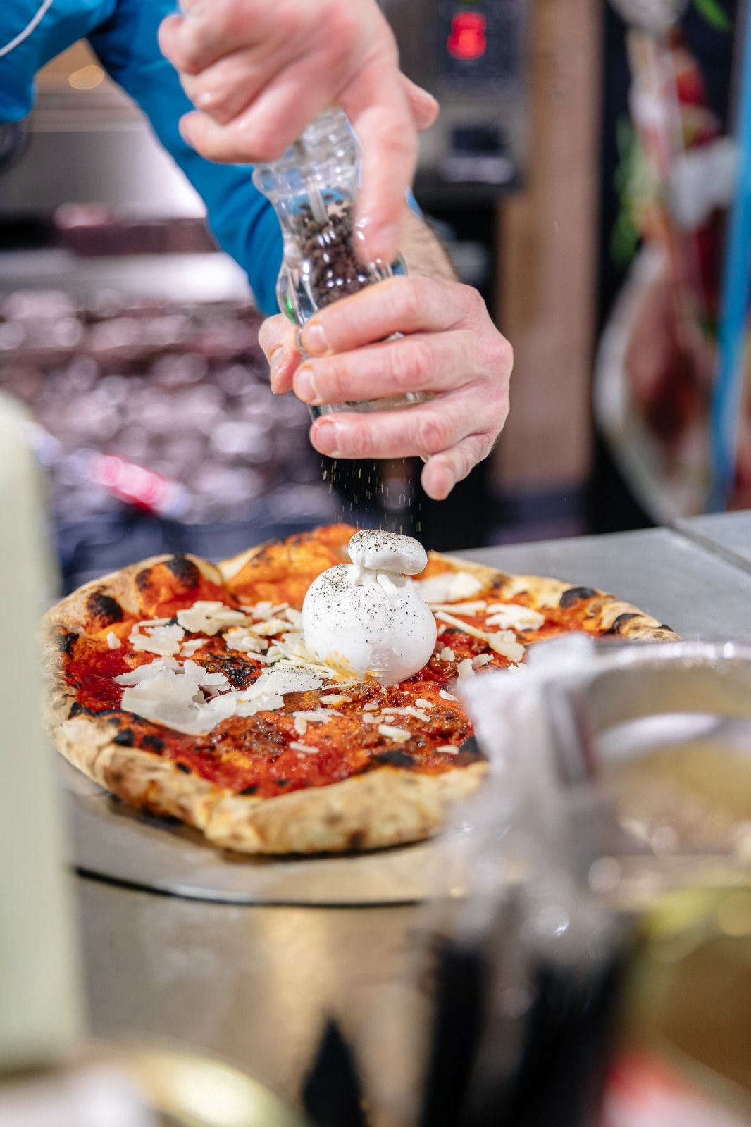 Chef seasoning a Neapolitan pizza topped with burrata and shaved cheese with a pepper grinder.
