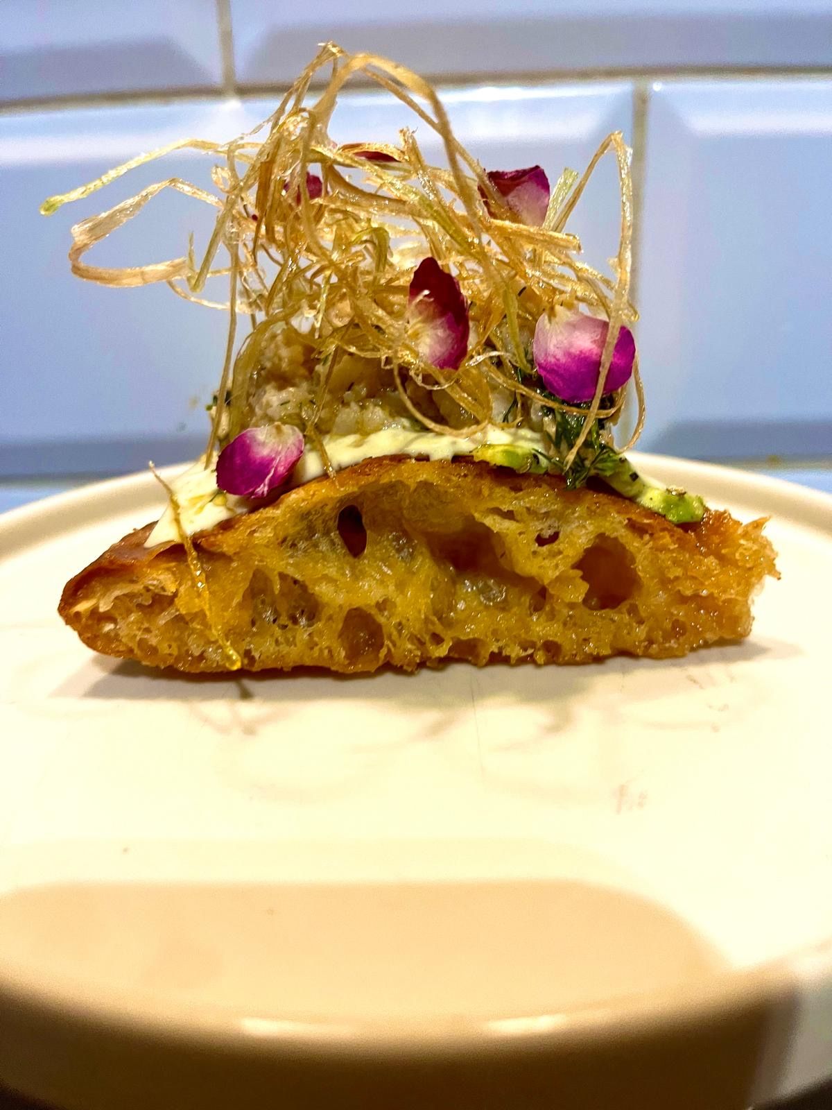 An artisan focaccia slice topped with avocado, edible flowers, and crispy fried leeks on a white plate.