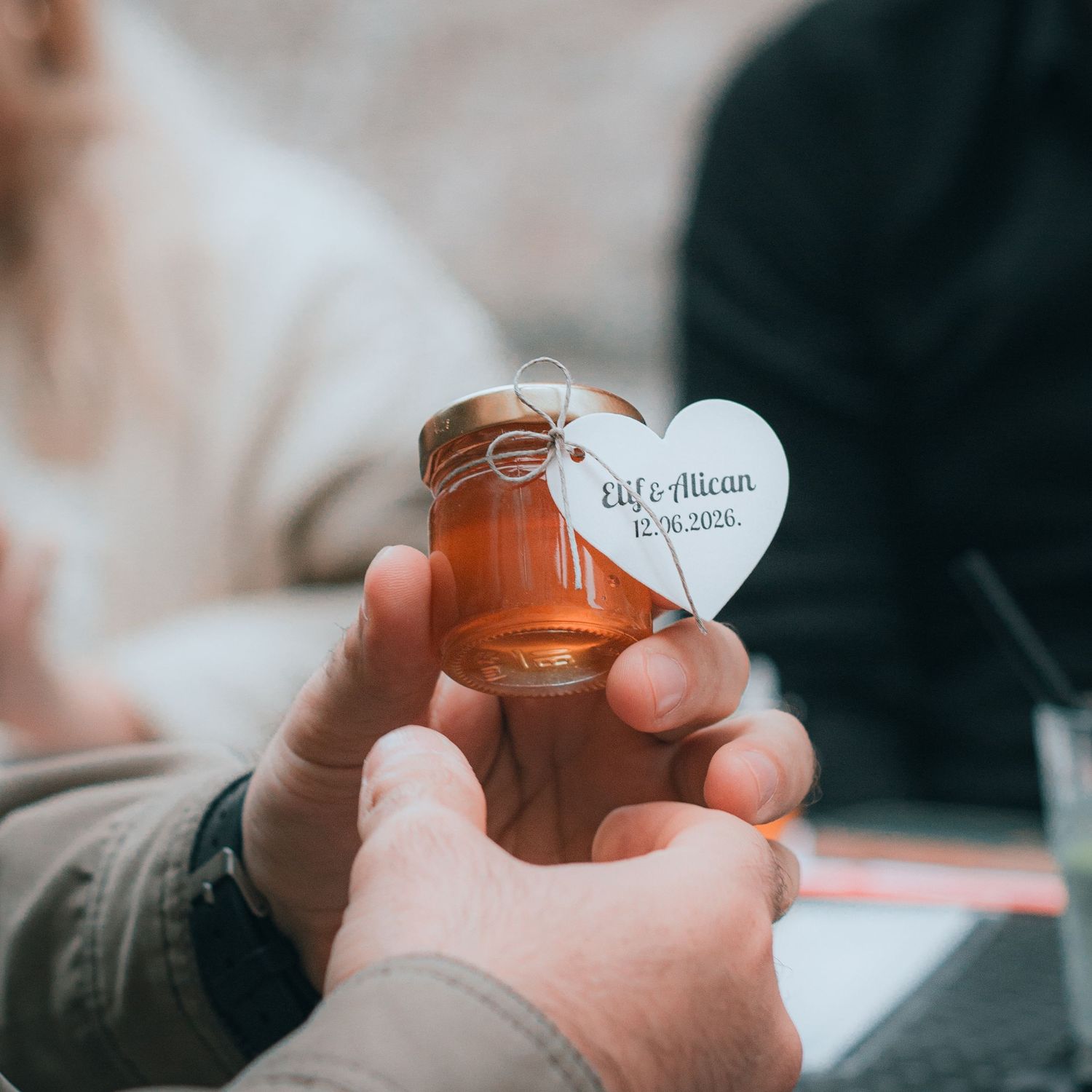 Small honey jar wedding favor with heart-shaped tag reading Elif & Alican 12.06.2020, held by hands.
