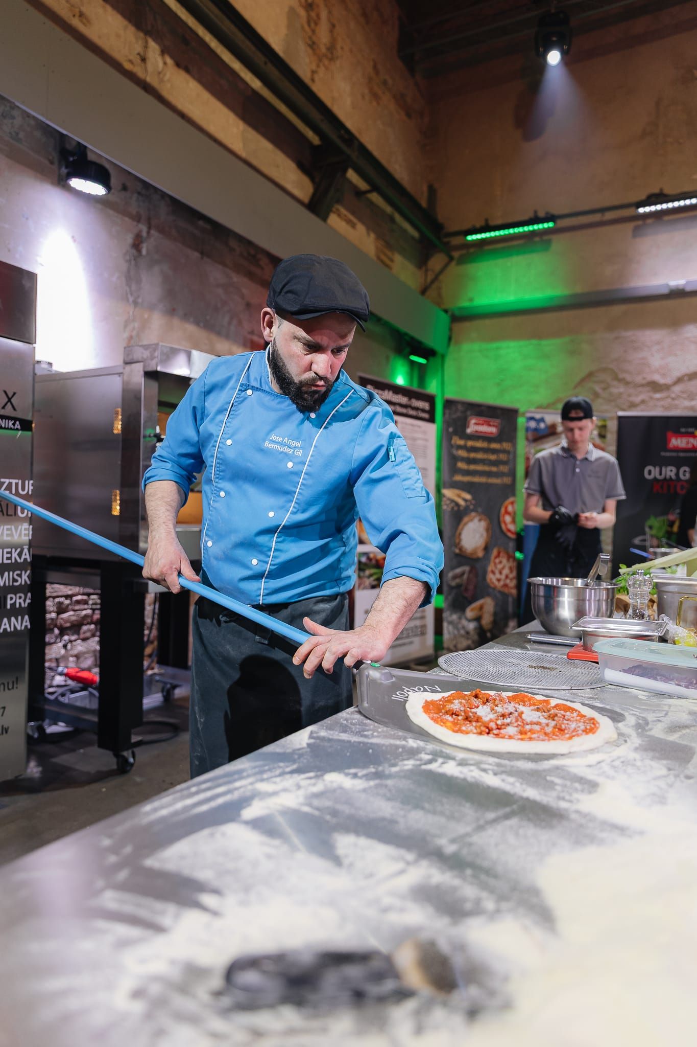 Male chef in blue uniform using a pizza peel to place a Margherita pizza into a competition oven.