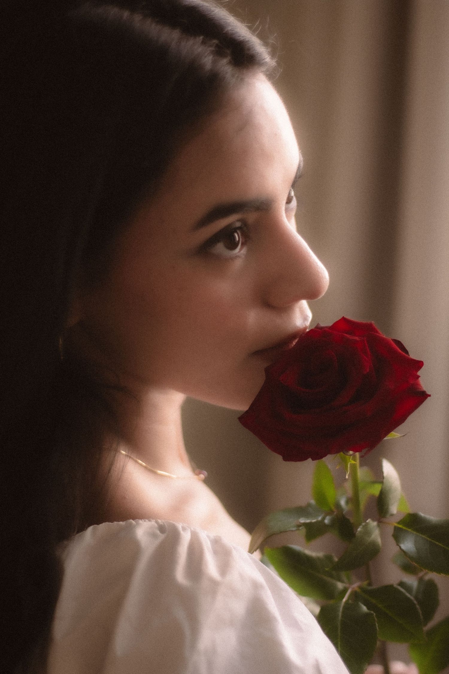 Young woman in white off-shoulder dress smelling a red rose in soft dramatic lighting.