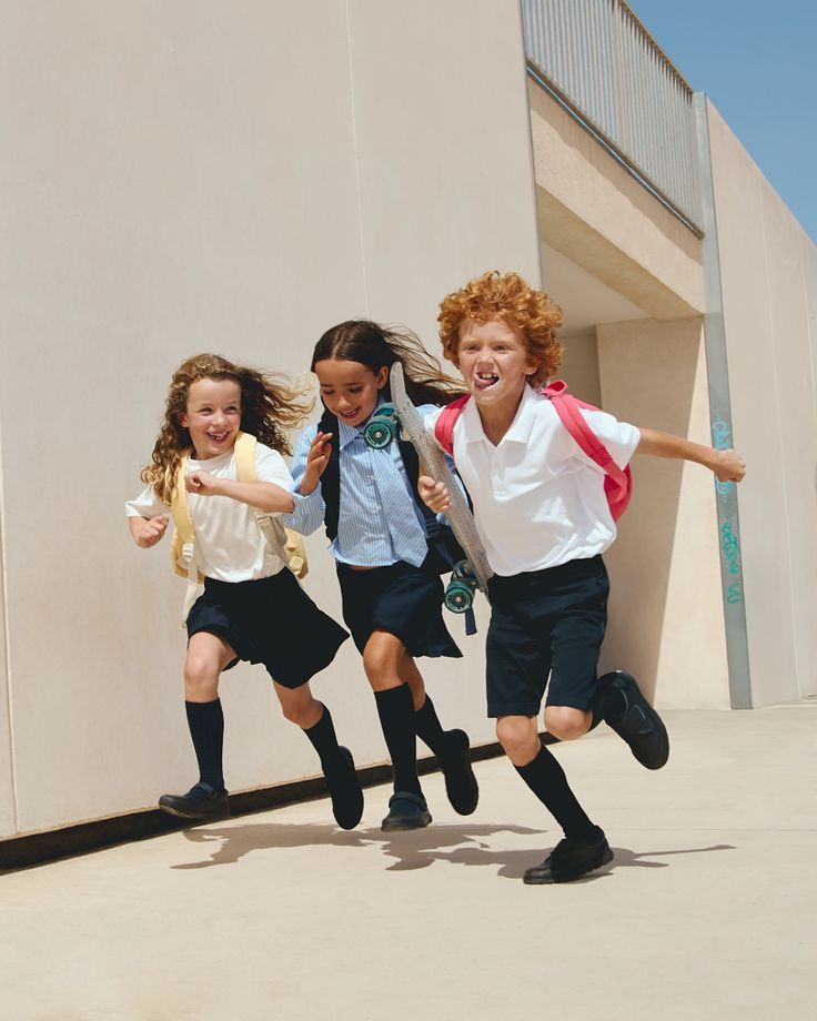 Three happy children in school uniforms running outdoors on a sunny day.