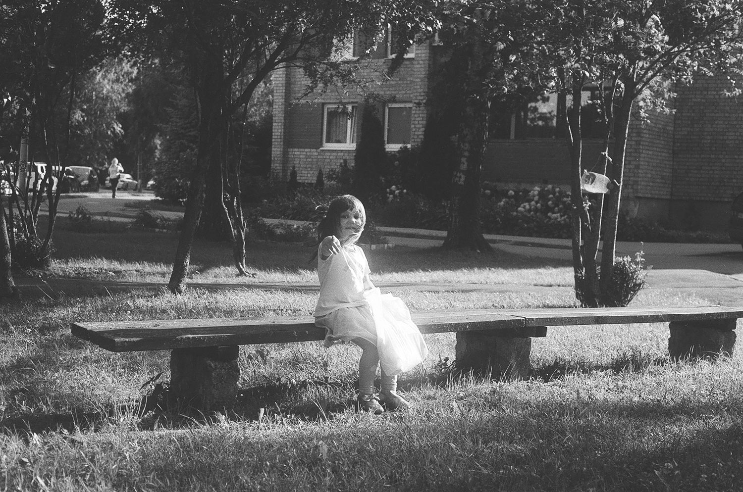 Black and white photo of a young girl in a white dress sitting alone on a stone bench in a park.