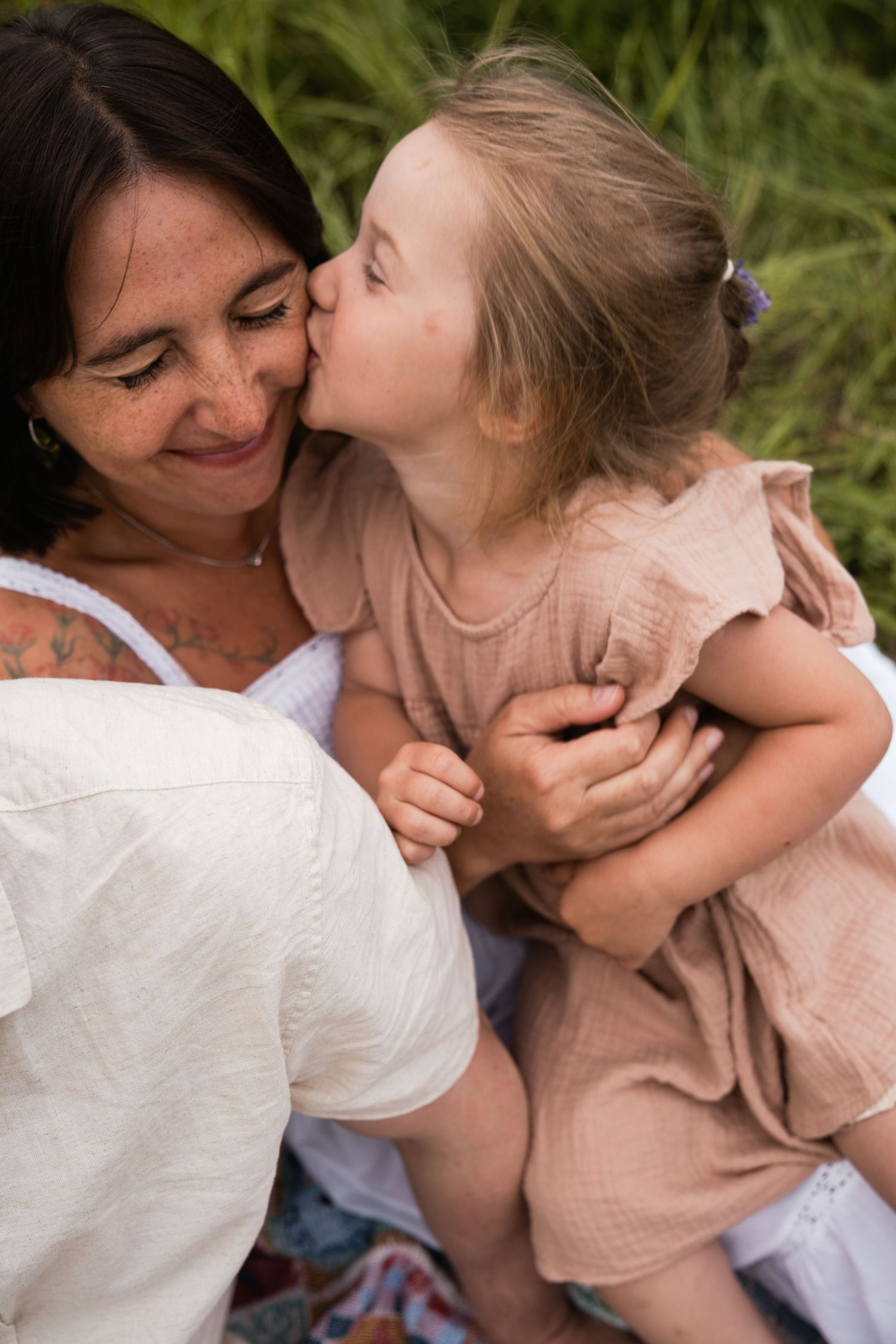 Young girl kissing her smiling mother on the cheek outdoors in a grassy field.