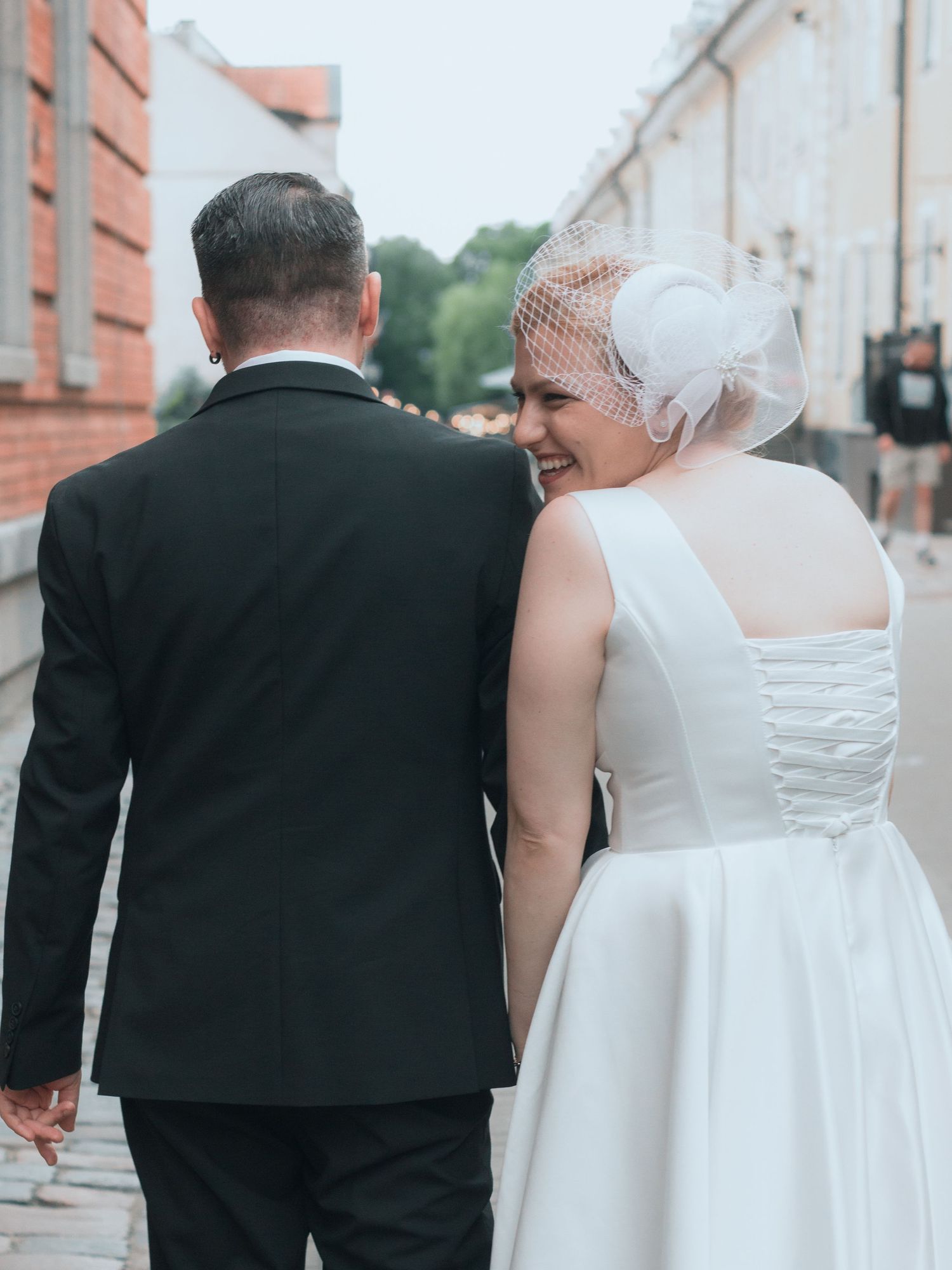A bride and groom walk hand in hand on a cobblestone street, she wears a white dress and birdcage veil.