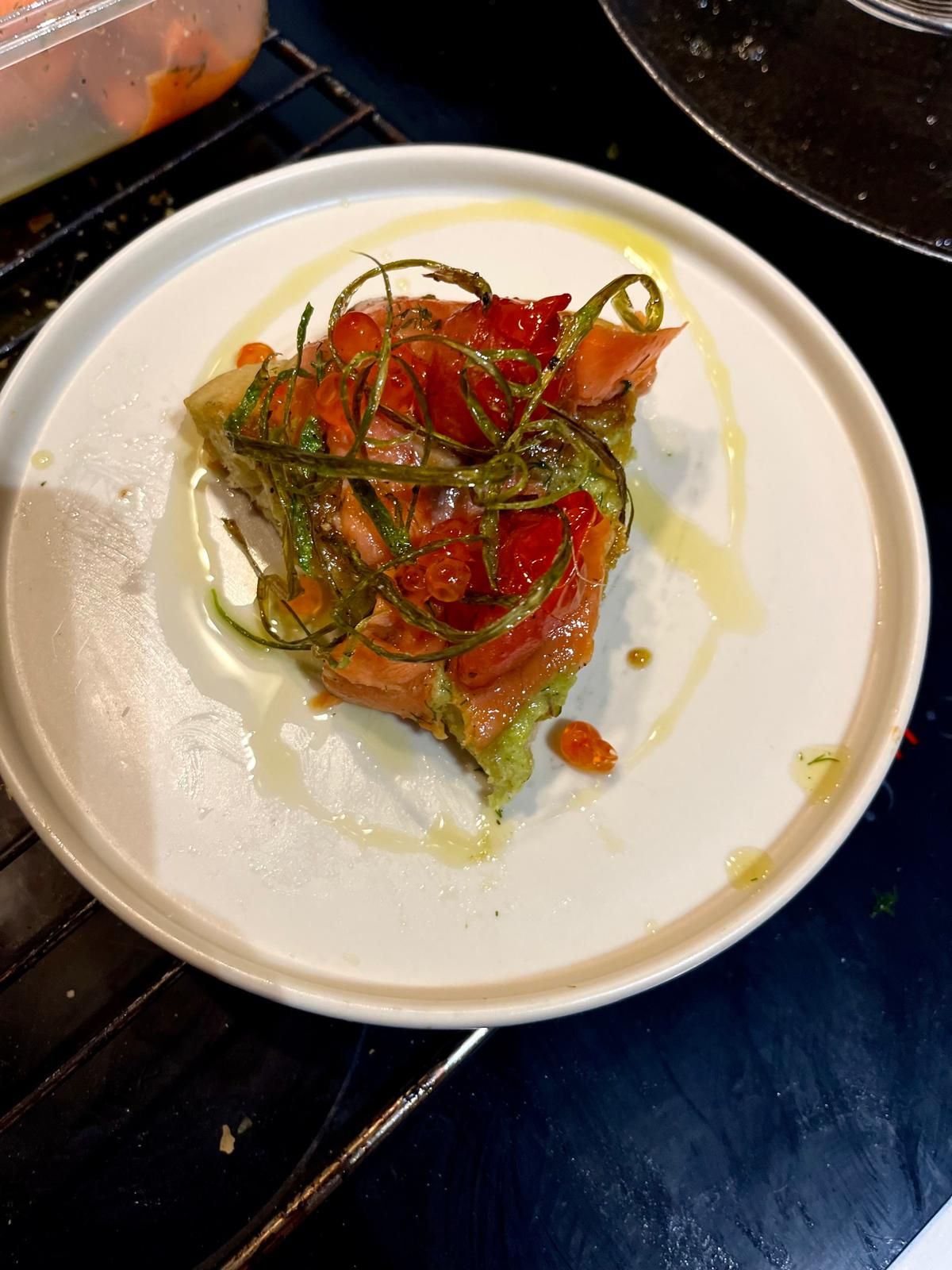 A plated focaccia topped with smoked salmon, roasted tomatoes, avocado, and herb garnish on white plate.