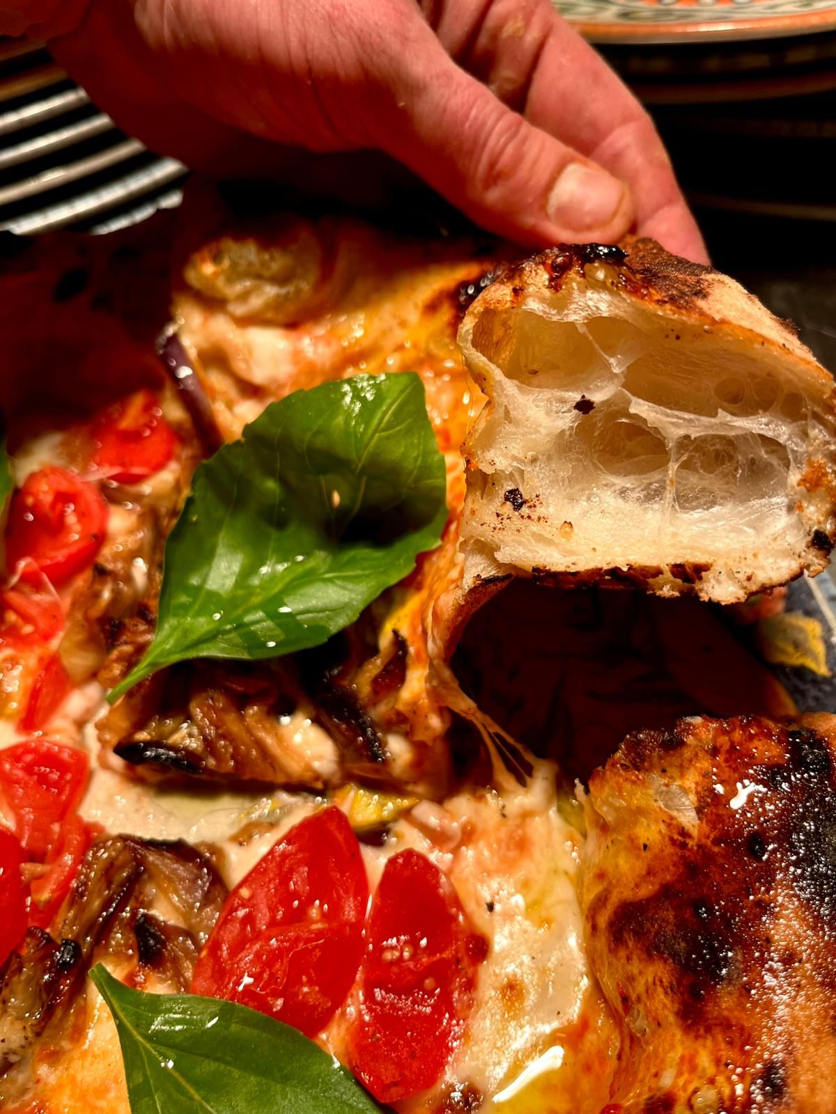Hand holding a piece of artisan pizza crust showing airy open crumb with tomatoes, mushrooms, and basil topping.