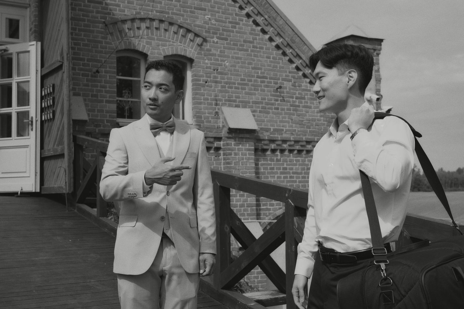 Groom in beige suit with bow tie chatting with guest outside brick building.