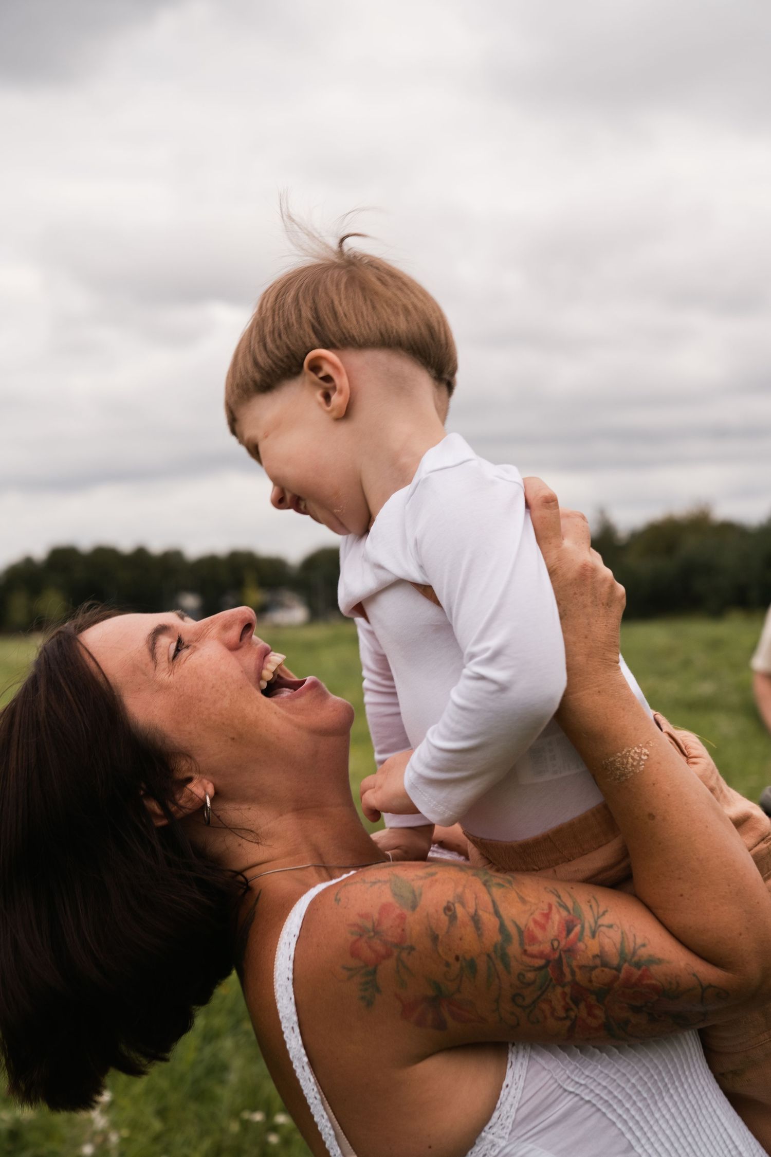 A tattooed mother joyfully lifts her laughing baby in a green field under a cloudy sky.