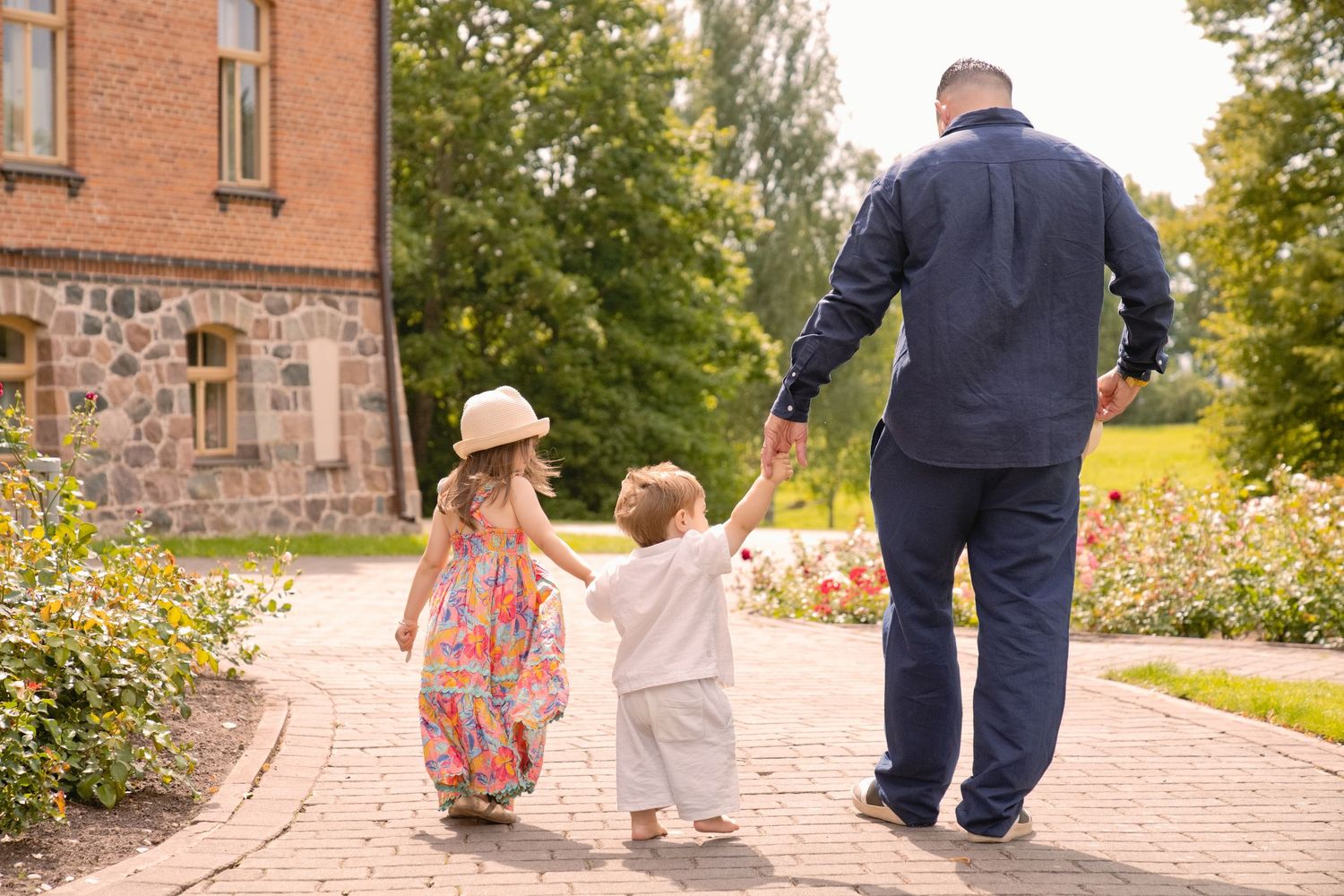 A father walks hand-in-hand with two young children along a brick path beside a stone building surrounded by greenery.