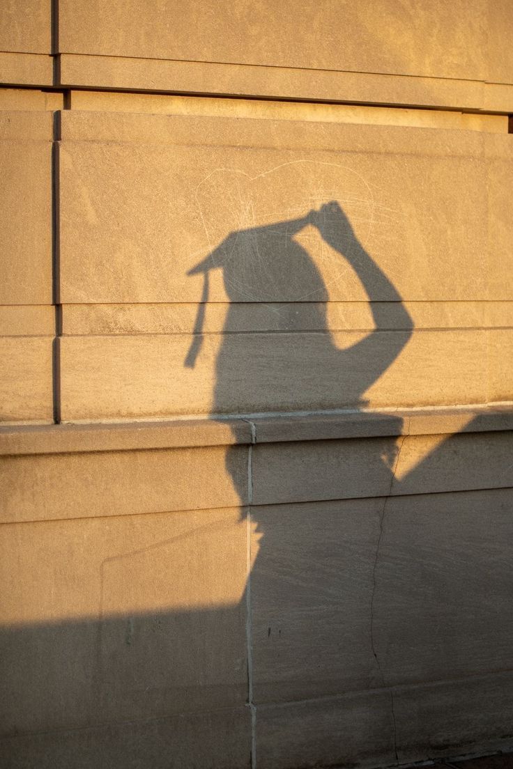 Shadow of a graduate in cap and gown cast on a golden stone wall in warm sunlight.