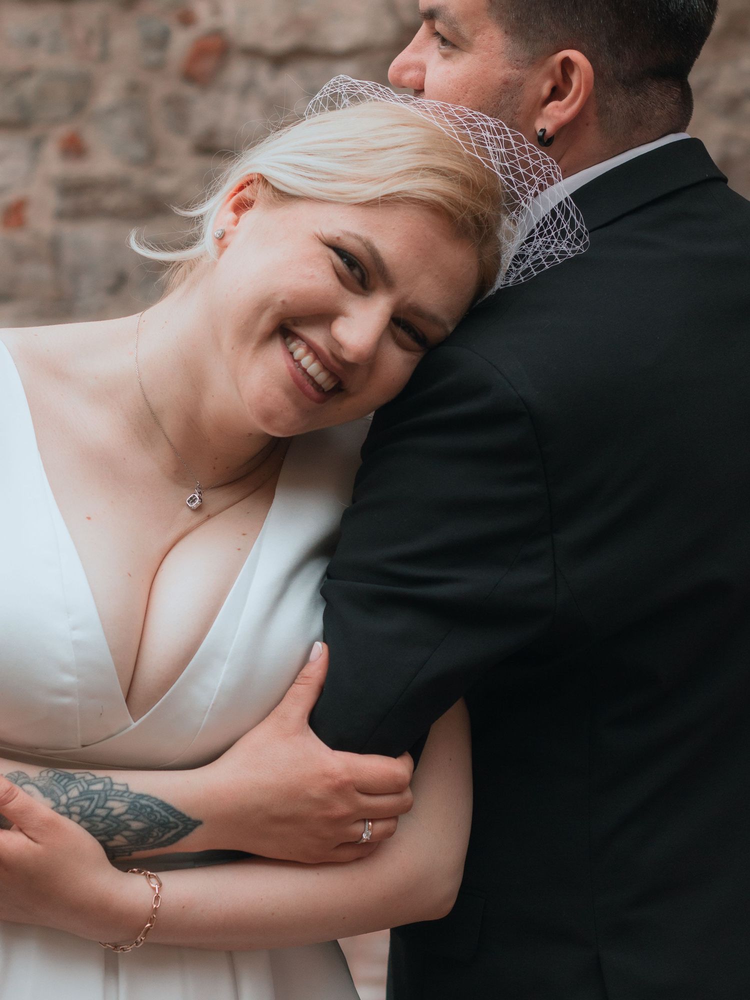 Smiling bride in white dress with veil embraces groom in black suit against rustic brick wall.