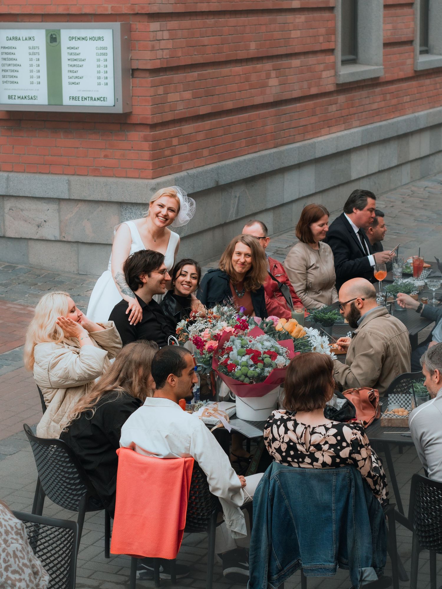 Bride in white dress stands smiling behind wedding guests seated at outdoor table with colorful floral centerpiece.