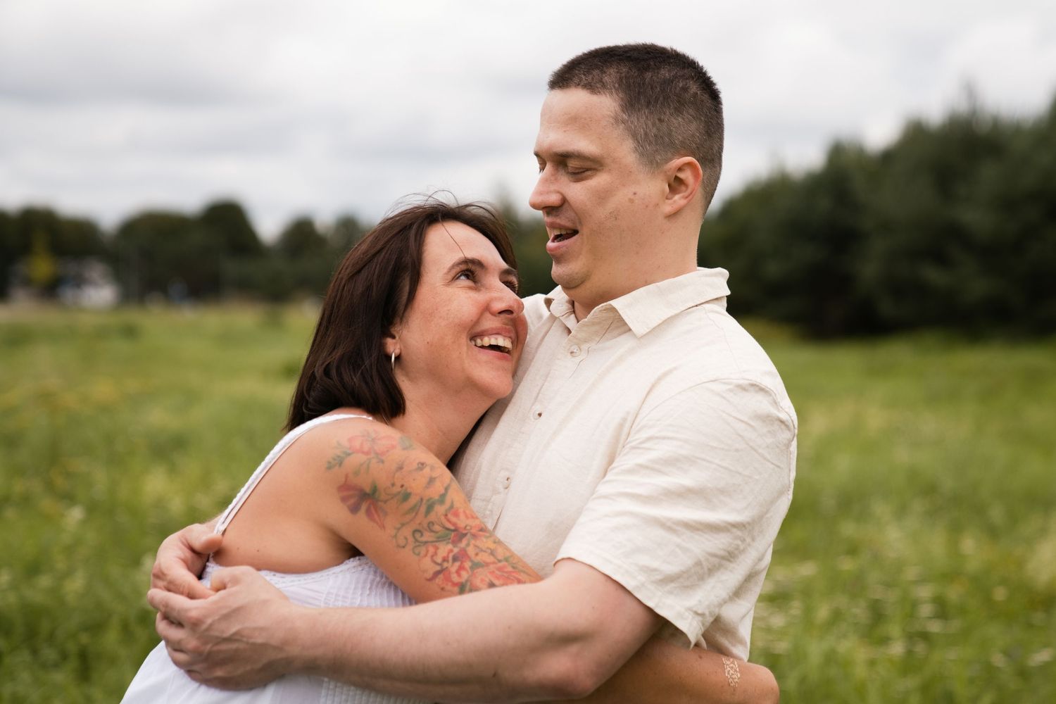 A smiling couple embraces in a green field, the woman with a floral arm tattoo laughing joyfully at her partner.