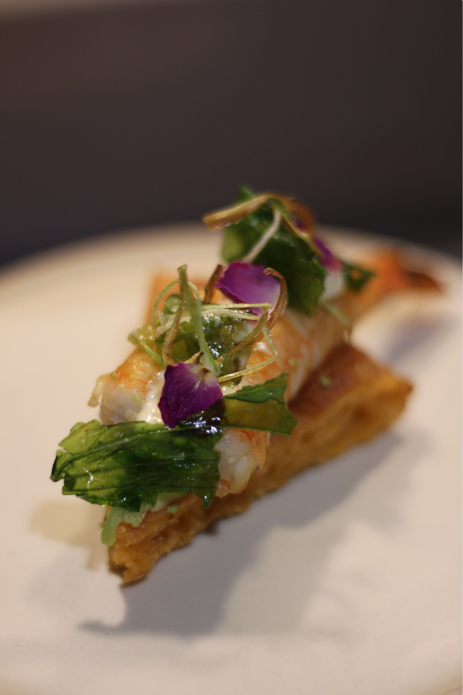 Elegant gourmet pizza slice topped with seafood, purple edible flowers, greens, and microgreens on white plate.