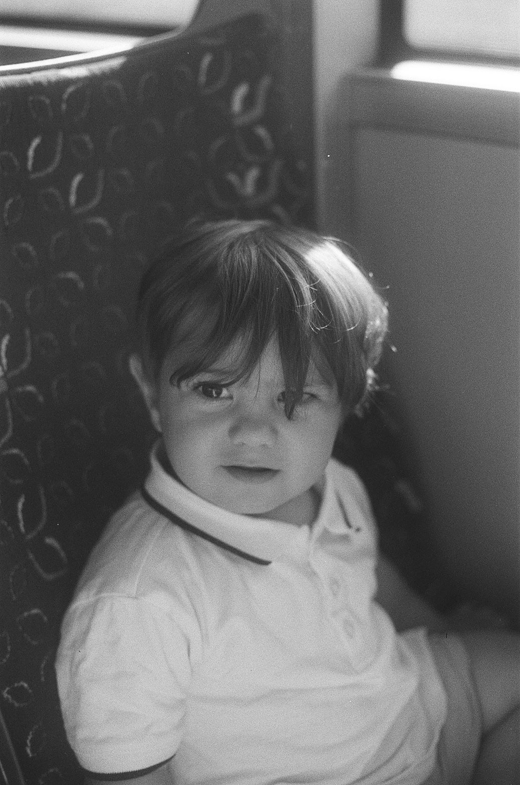 Black and white photo of a young toddler with short hair sitting in a patterned chair, wearing a white polo shirt.