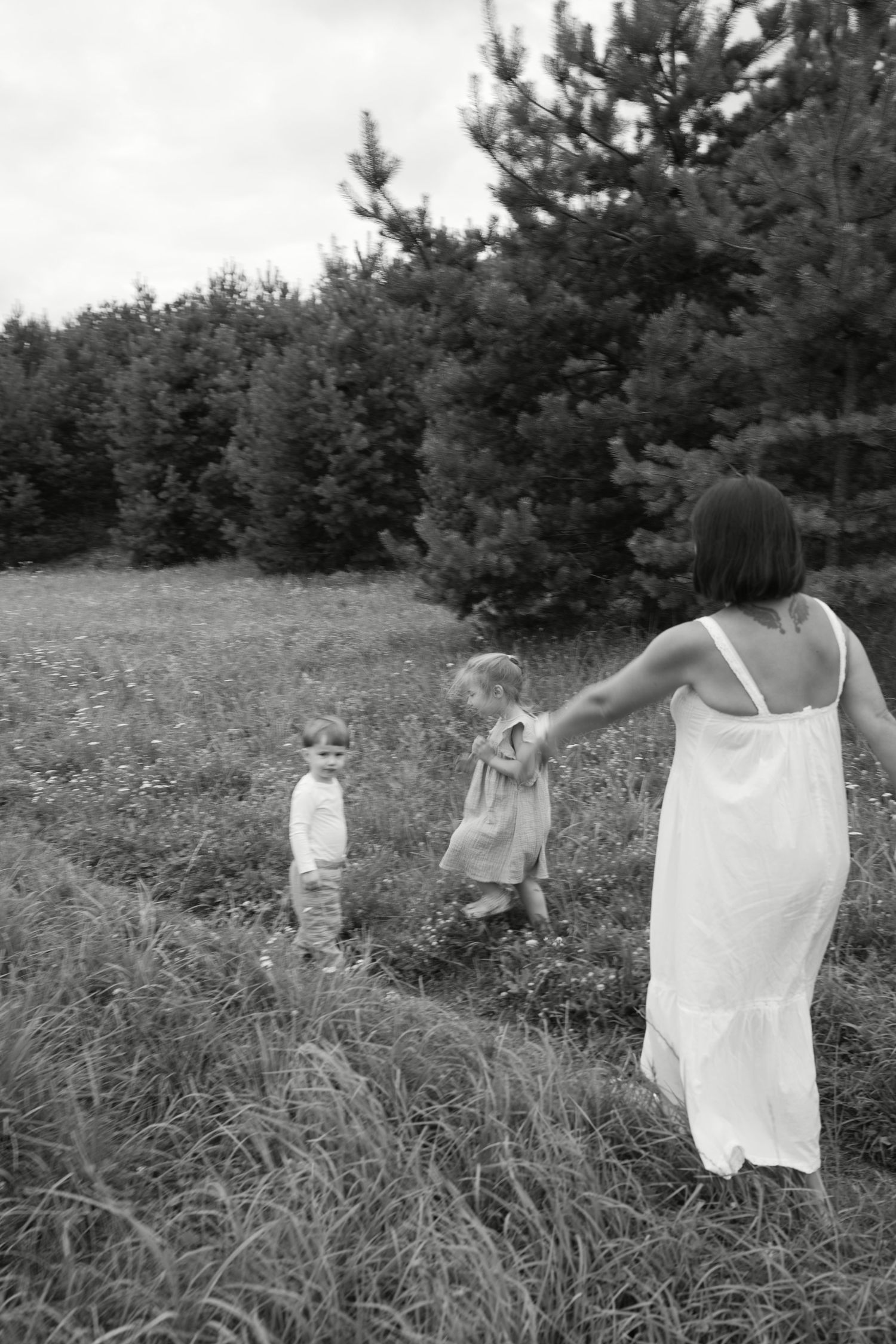 Black and white photo of a mother in white dress holding hands with two young children in a grassy field near pine trees.