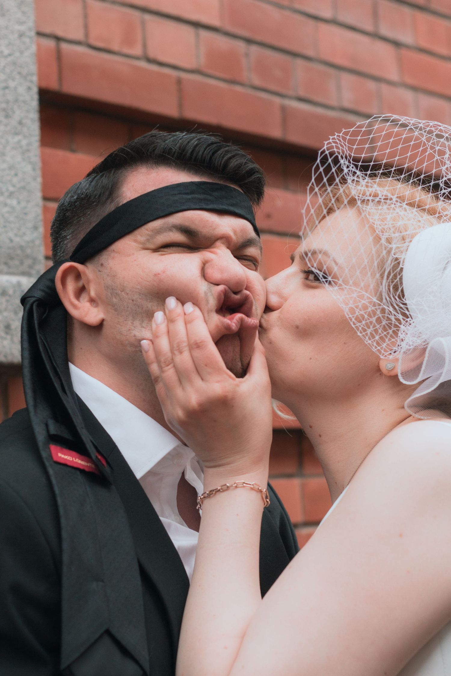 Playful bride in white veil kisses groom wearing black blindfold tie at outdoor wedding celebration.