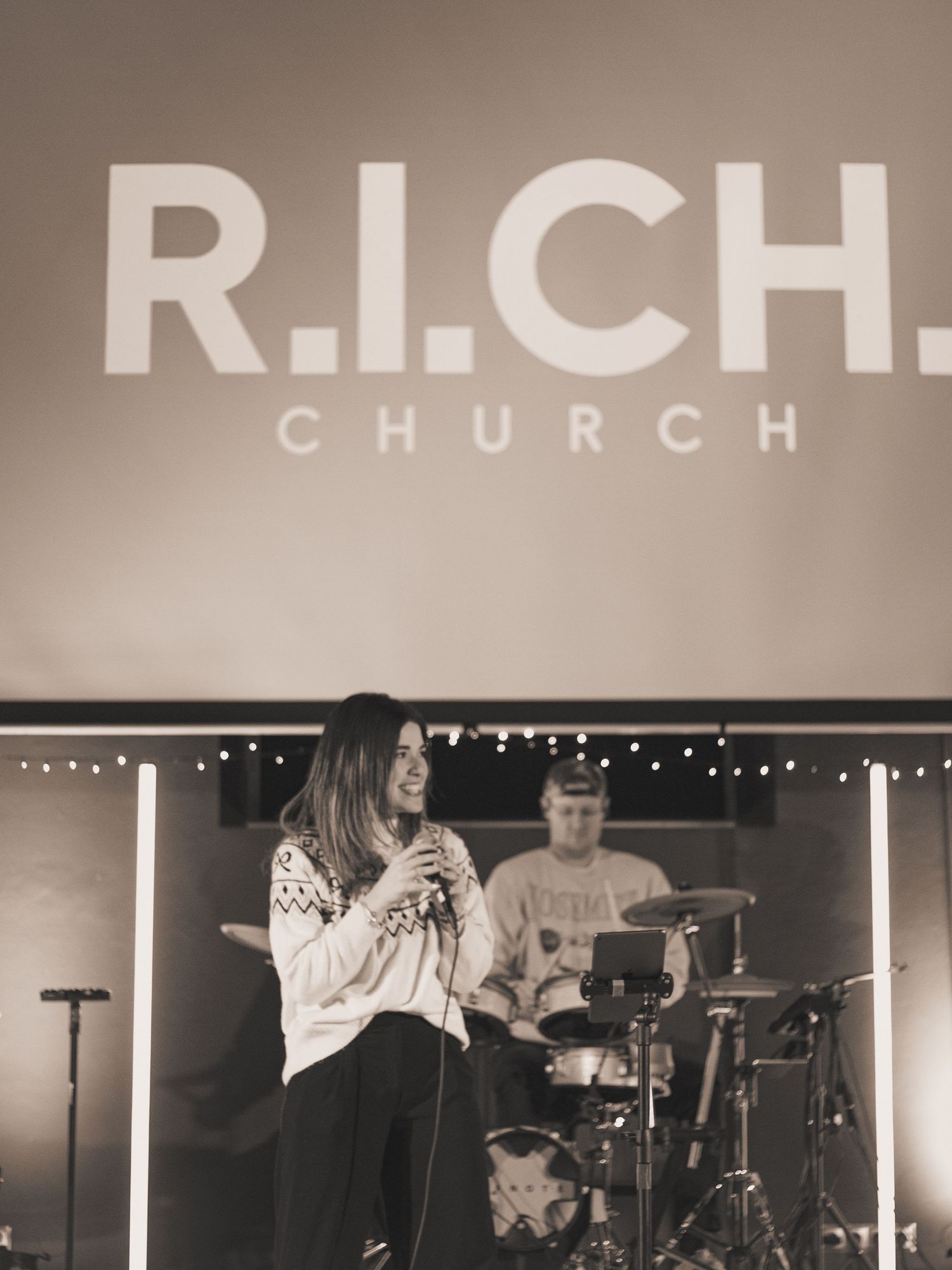 Black and White image of a woman singing and a drummer behind her. The words 