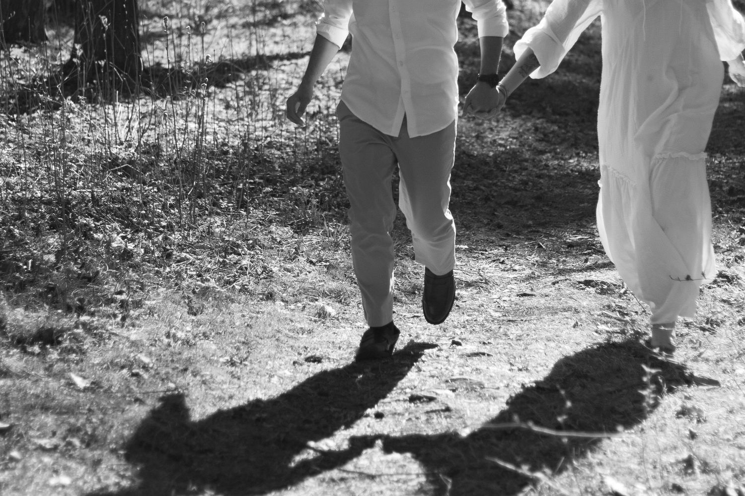 Black and white photo of couple walking on forest path, casting shadows on ground in sunlight.