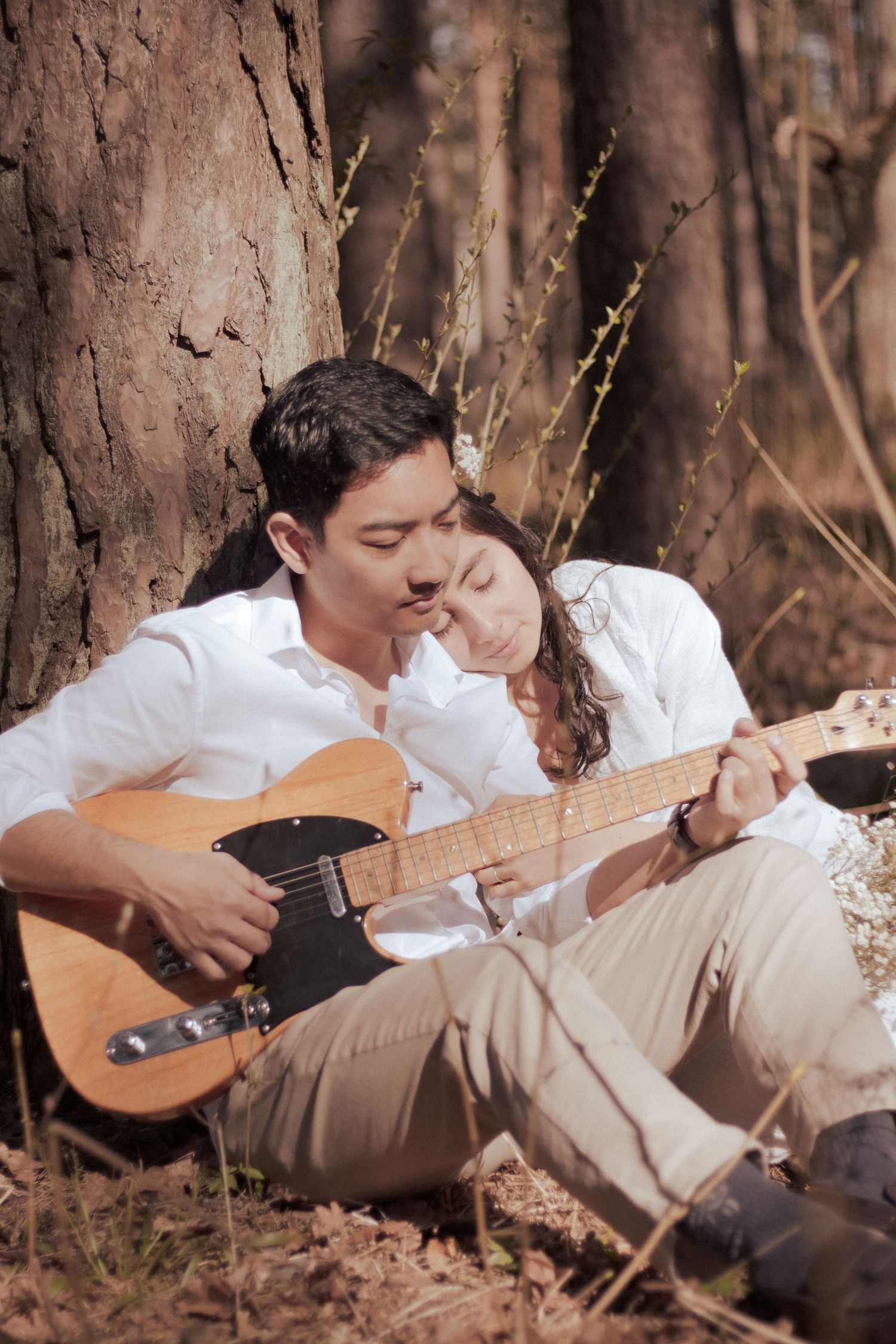 Man playing electric guitar with woman resting on his shoulder by forest tree.