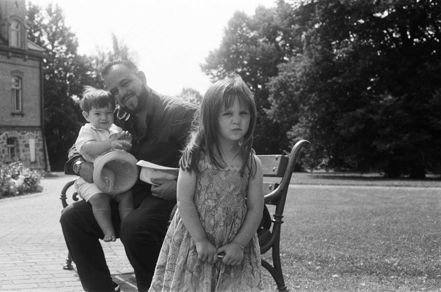 Father sitting on bench with two young children in sunlit park, black and white.