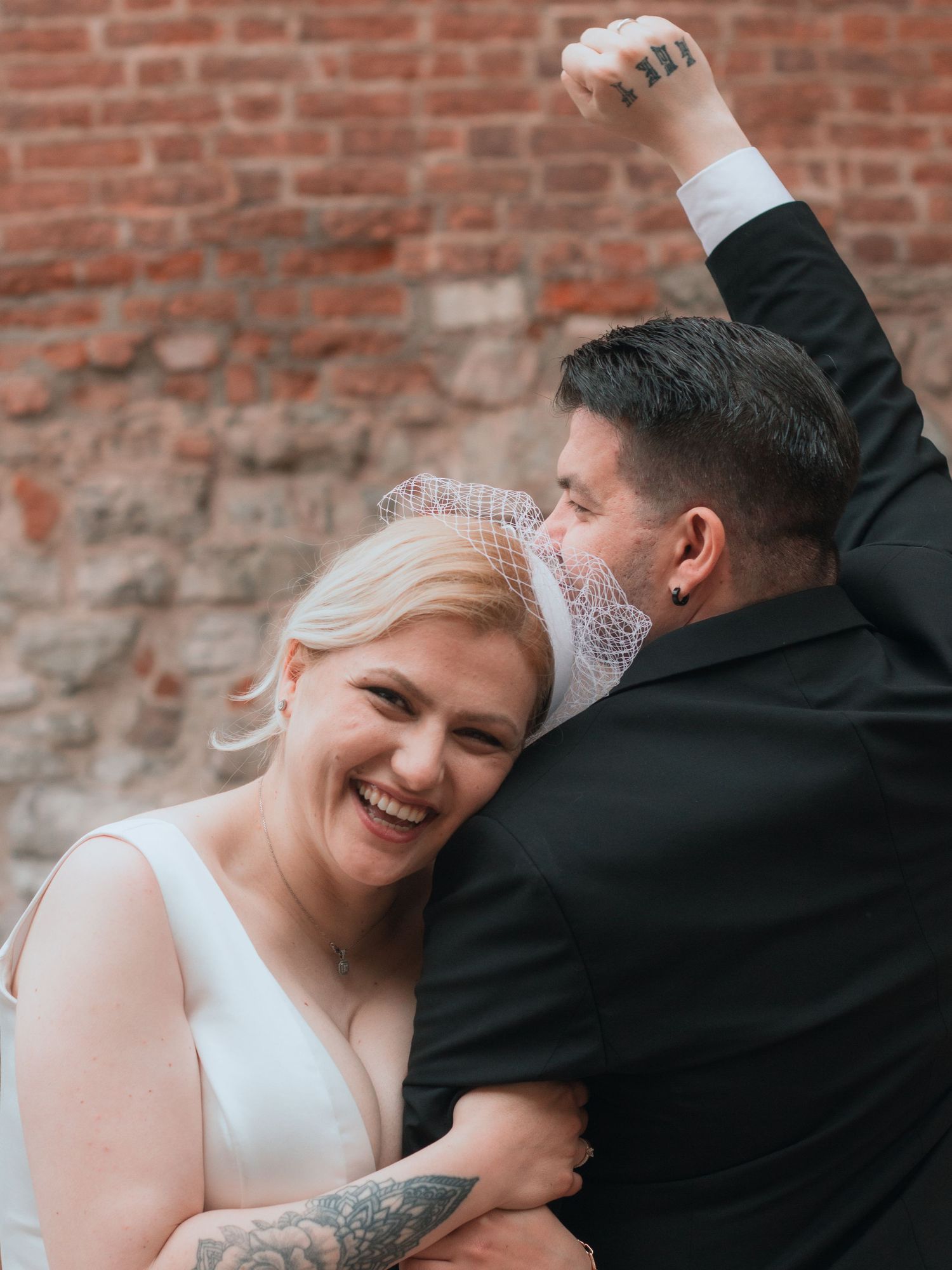 Joyful bride in white dress and veil laughs as groom in black suit raises fist in celebration against brick wall.