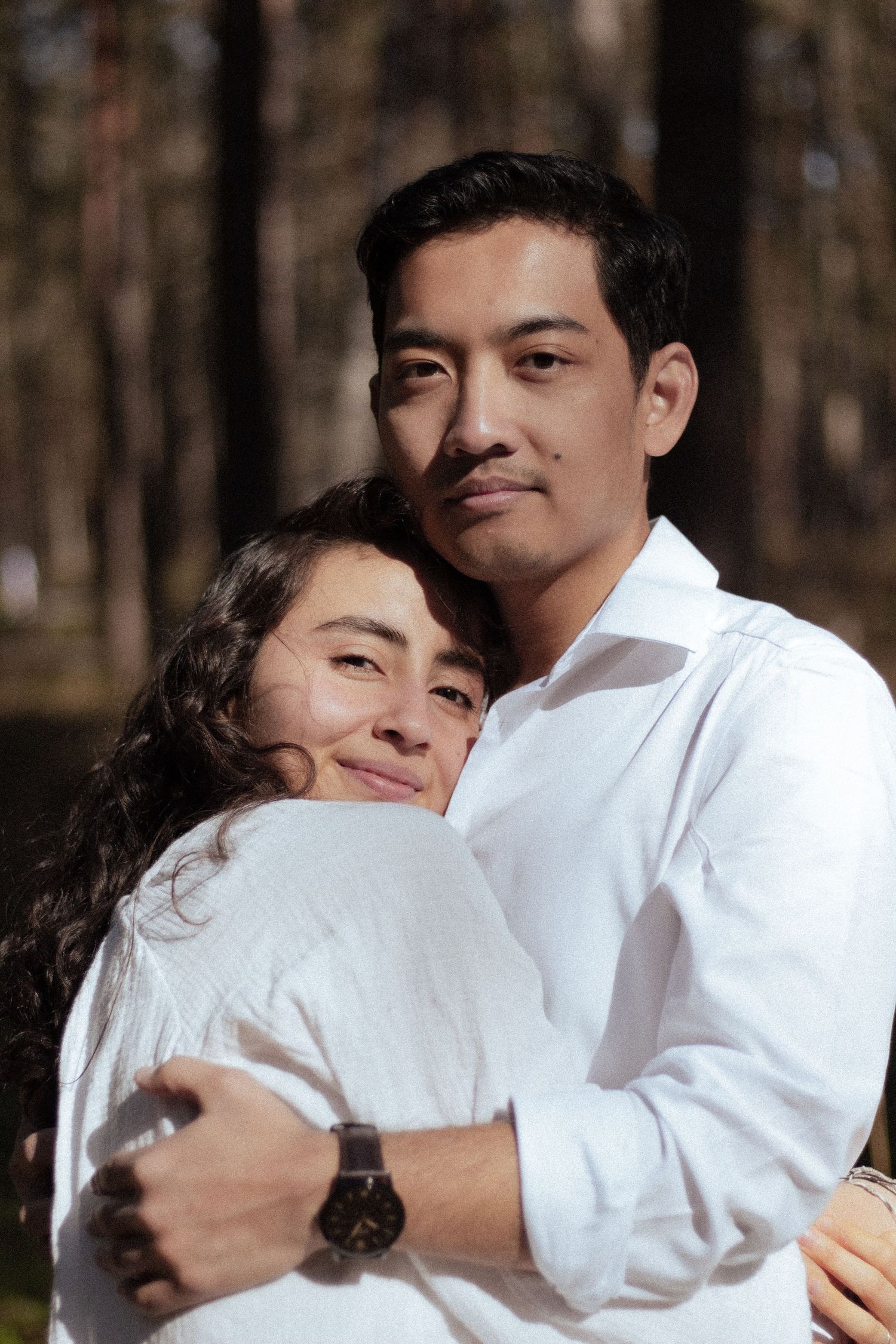 Couple embracing outdoors in white shirts with sunlit pine forest background.