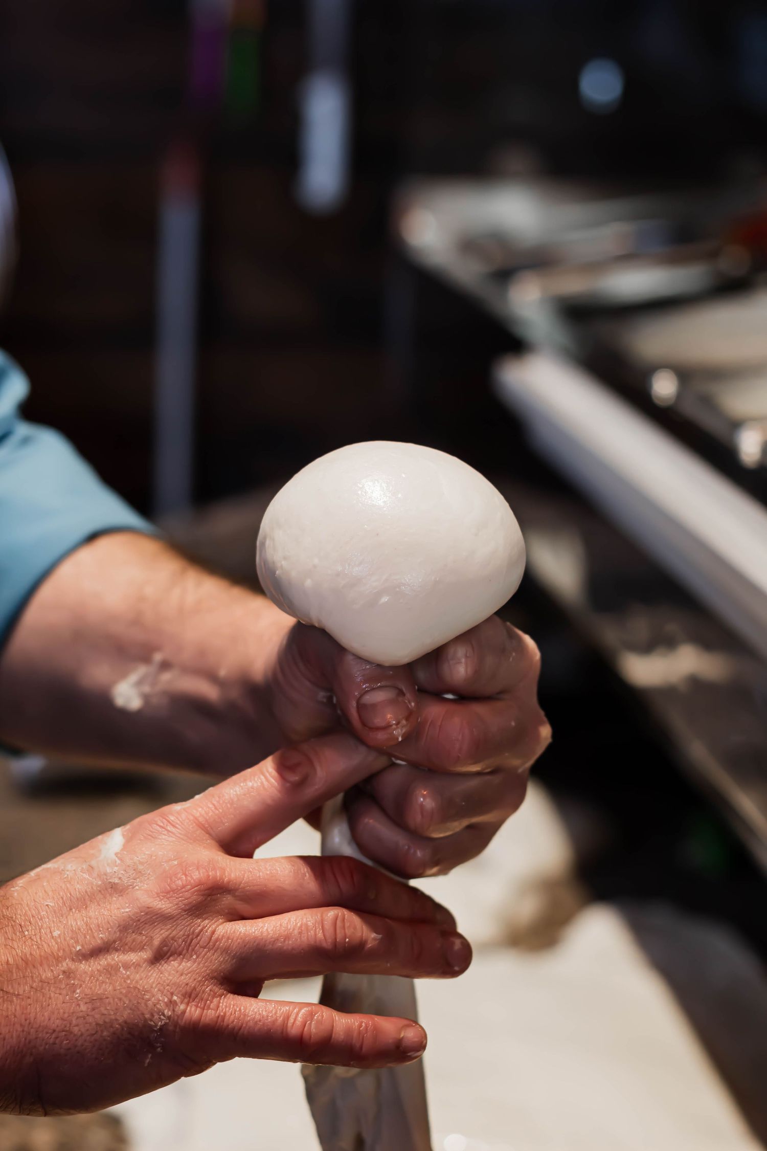 Chef's hands shaping a ball of fresh mozzarella cheese over a kitchen counter.