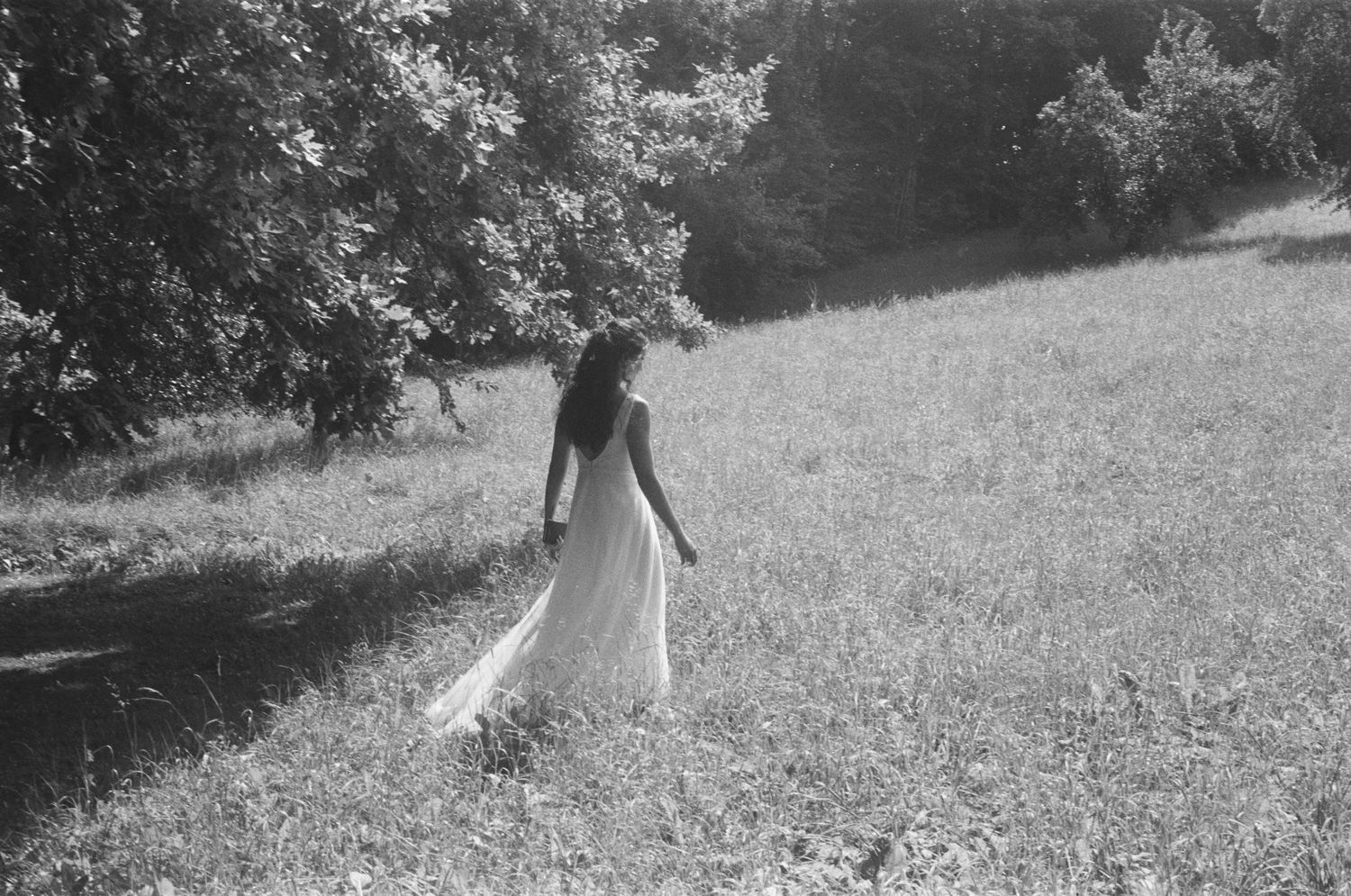 Bride in flowing white gown walking through sunlit meadow toward trees, black and white.