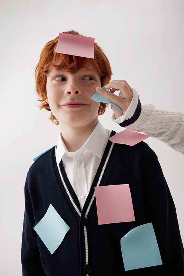 Redhead boy with colorful sticky notes on his face and uniform in classroom.