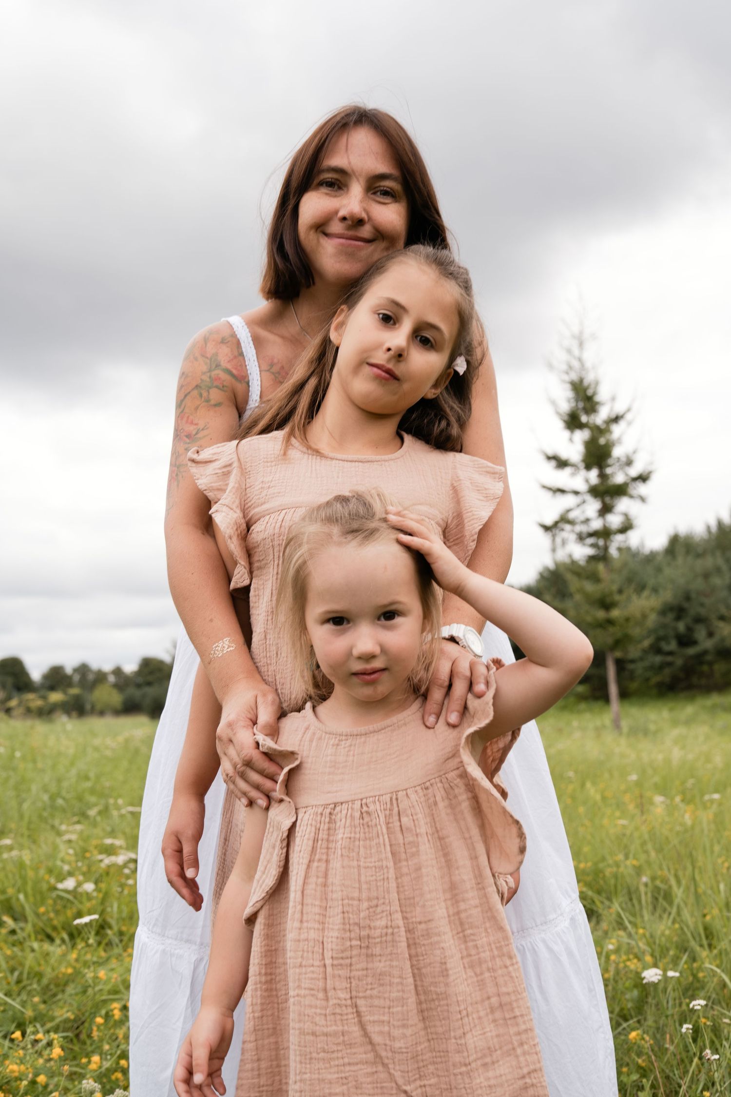 A mother and two daughters in matching earth-tone dresses pose together in a green meadow under a cloudy sky.