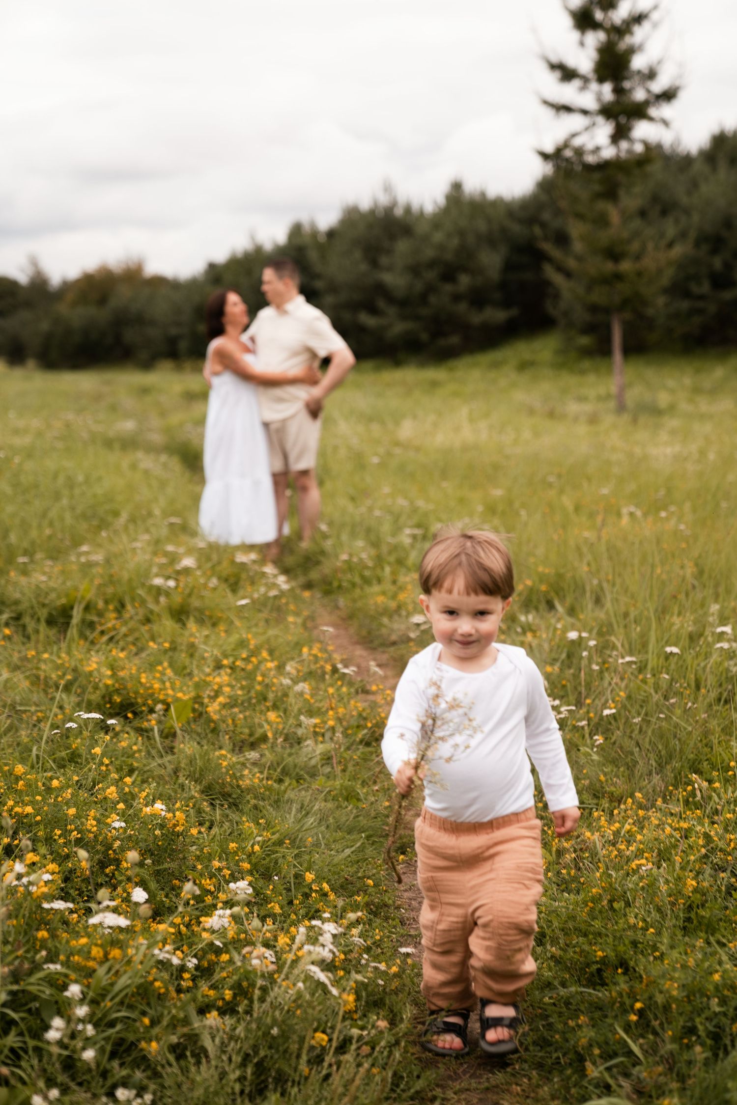 A young child holds wildflowers in a meadow while parents embrace on a path surrounded by green fields and trees.