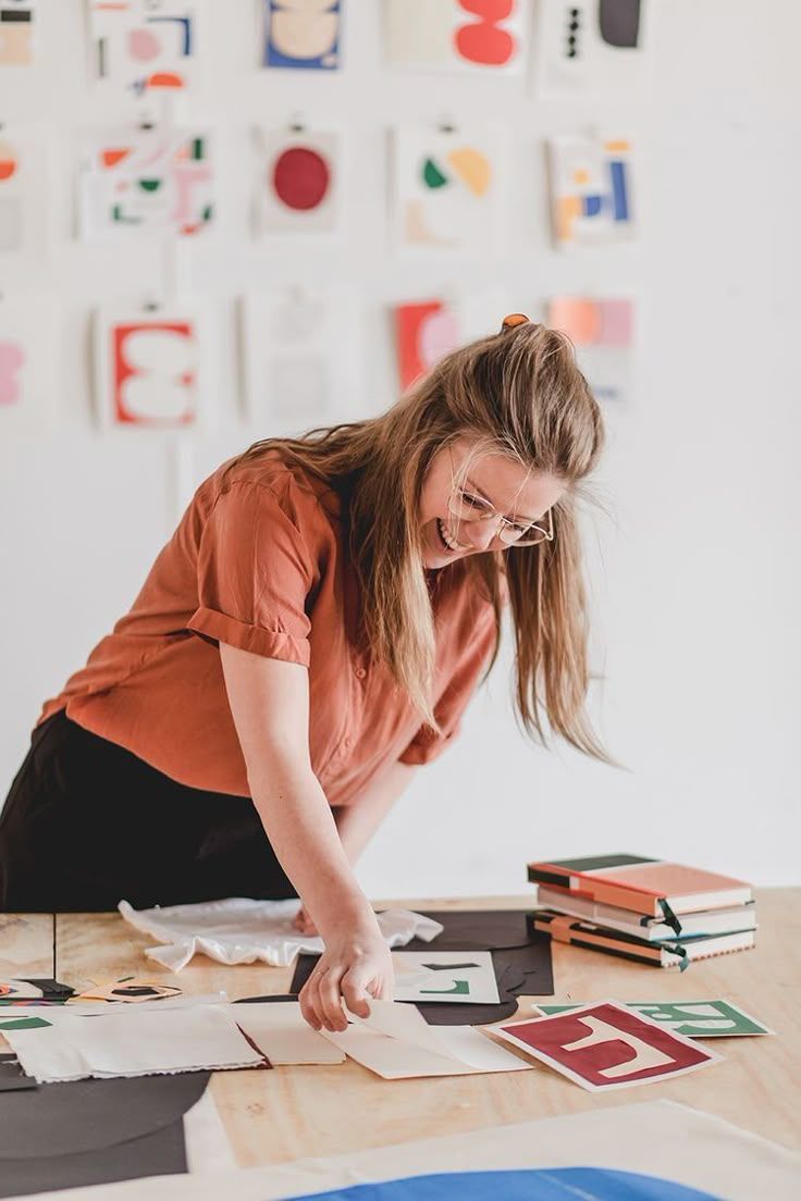 Smiling woman in rust blouse arranging colorful art prints on a wooden table in a creative studio.