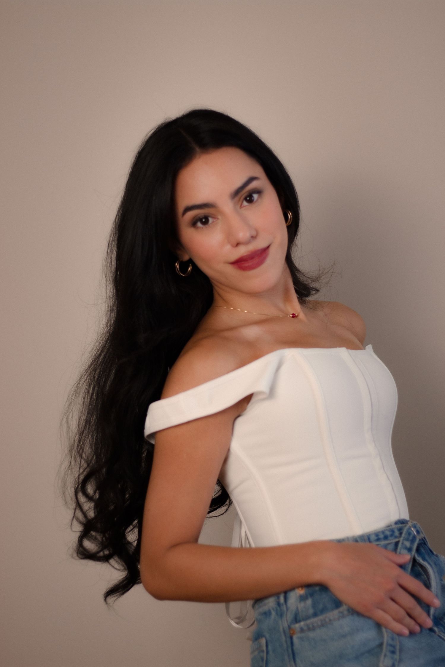 Young woman with long black hair in white off-shoulder corset top and gold jewelry, studio portrait.