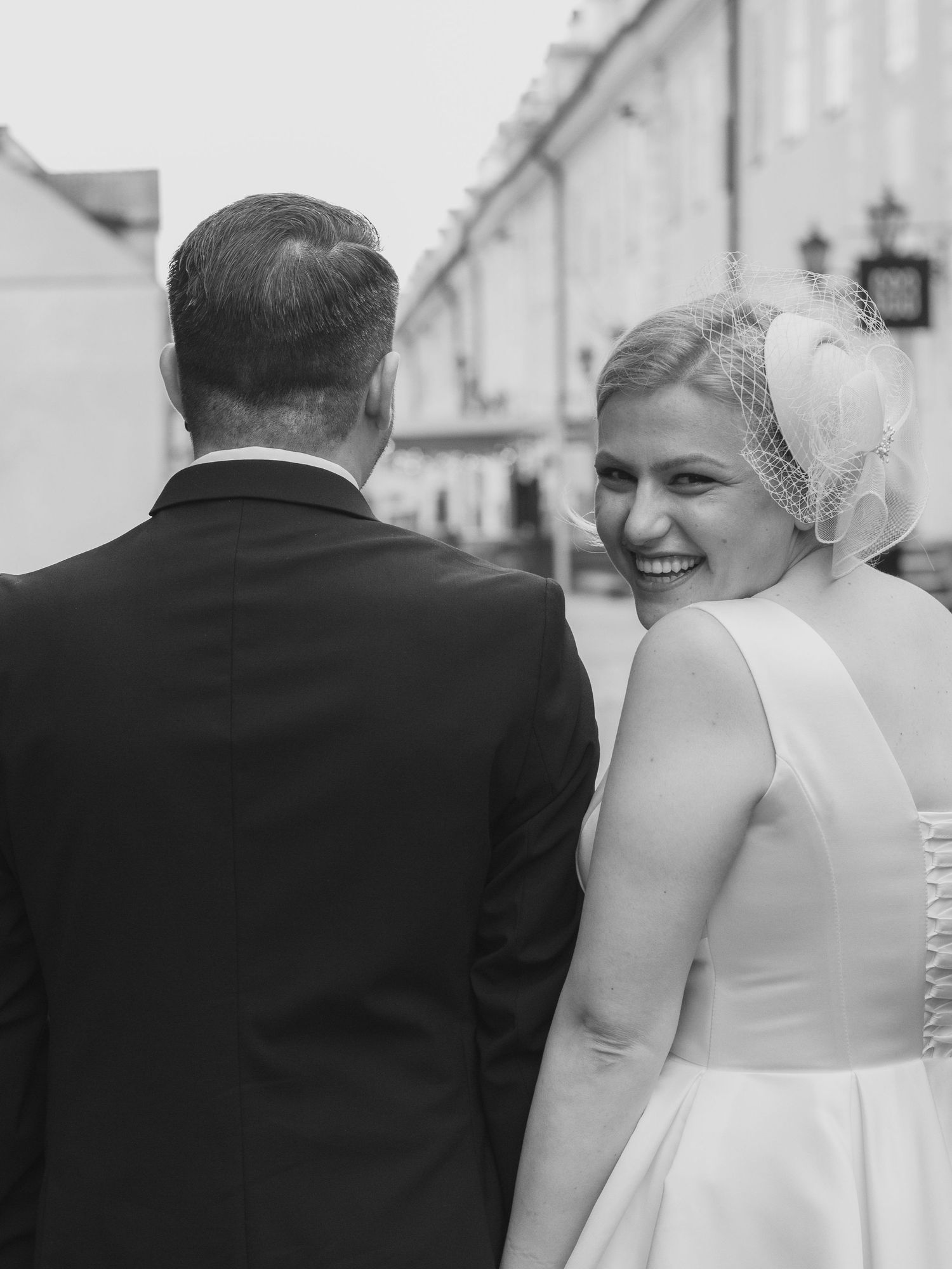 Smiling bride in white dress and birdcage veil walks with groom in black suit on a cobblestone street.