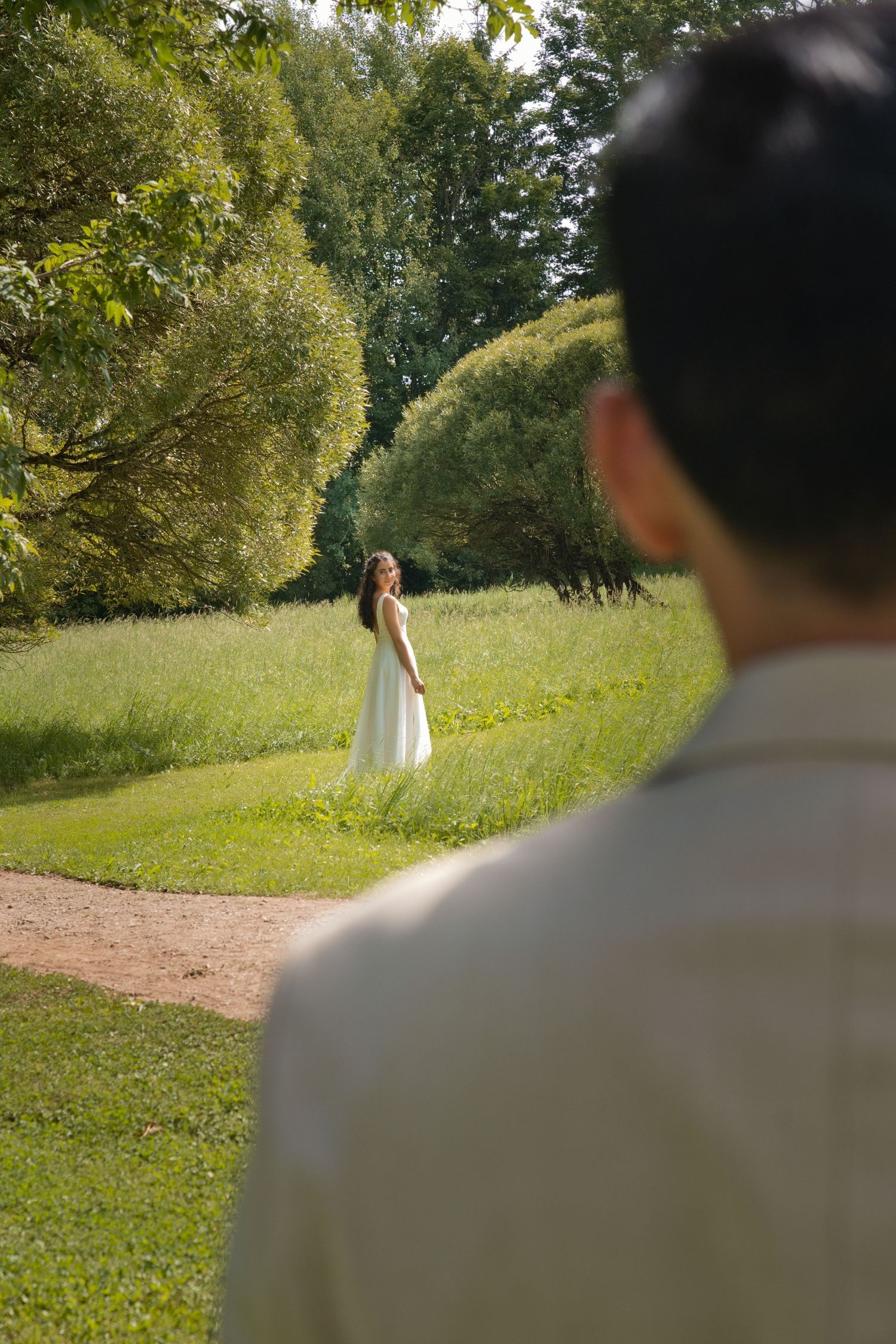 Bride in white dress standing in lush green park while groom watches from foreground.