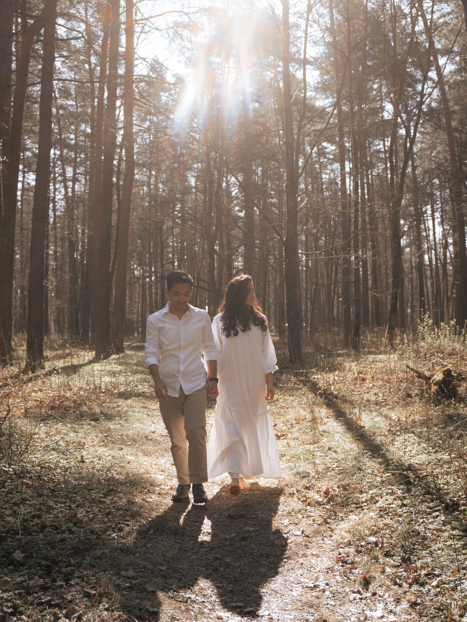 Couple holding hands walking through sunlit forest path, woman in white dress, man in white shirt, golden light streaming through trees.