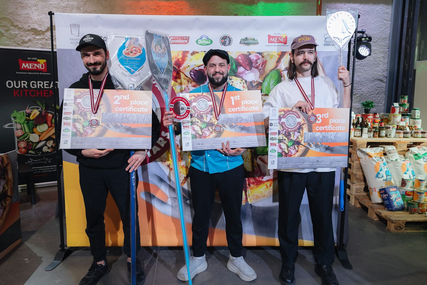 Three male pizza competition winners posing with medals and large prize boards on a stage.