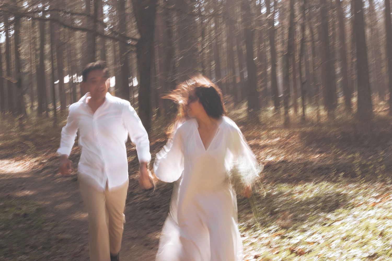 Couple in white outfits running hand in hand through a sunlit forest path.